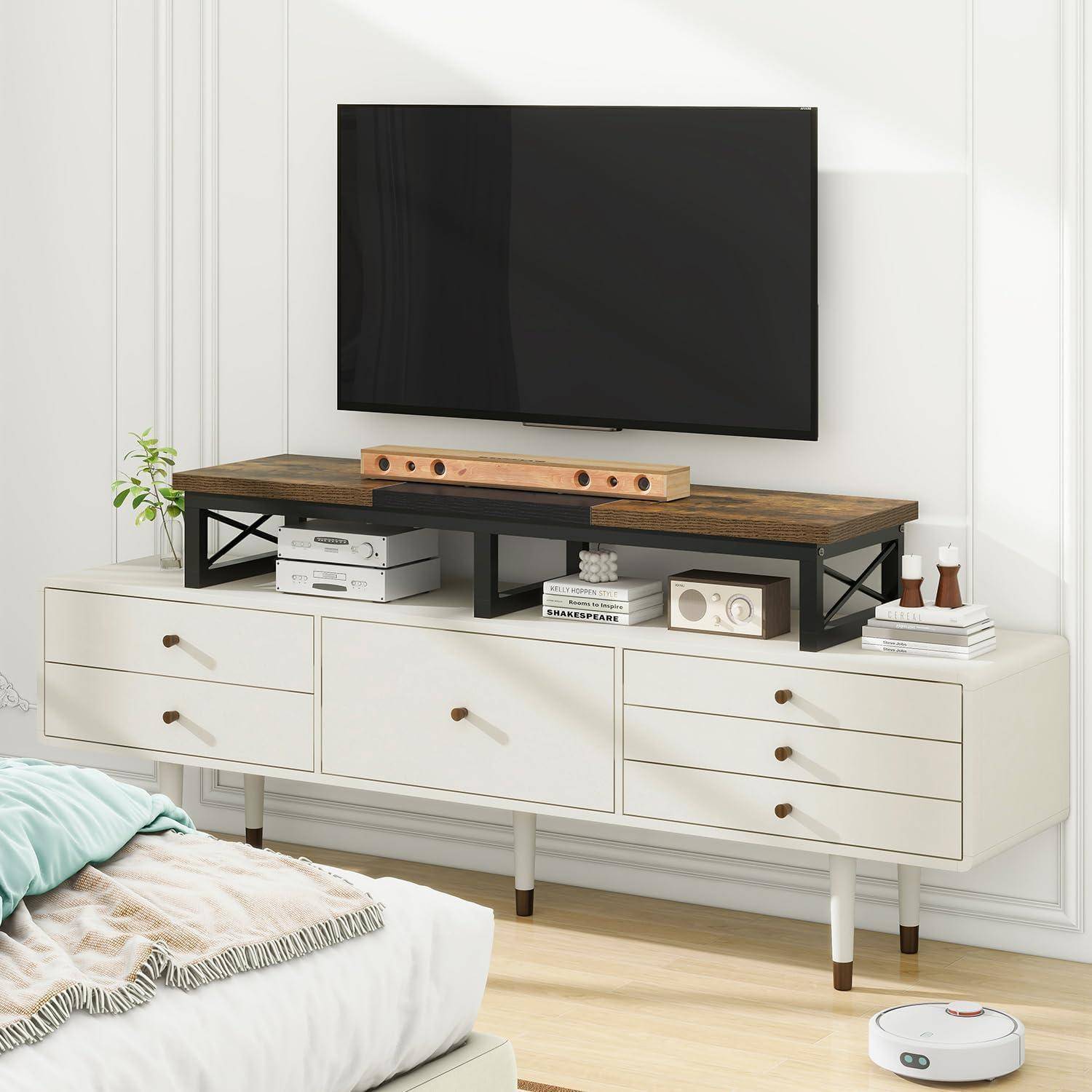Ein modernes Wohnzimmer zeigt einen an der Wand montierten Fernseher über einer weißen Konsole. Die Konsole trägt Bücher und ein Holzregal. Ein Bett und ein Roboterstaubsauger sind zu sehen.