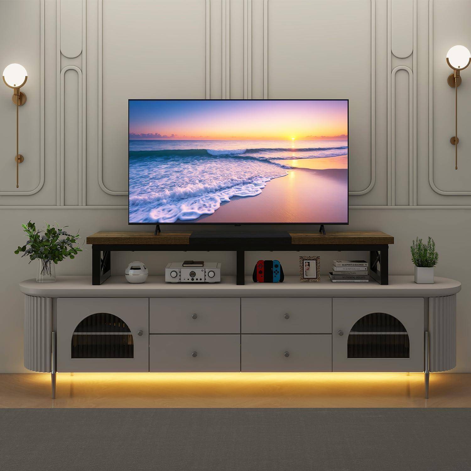 Wohnzimmer mit einem Fernseher, der einen Strand-Sonnenuntergang auf einem Holzständer über einer weißen Konsole zeigt. Pflanzen und Wandleuchten sorgen für die Dekoration.