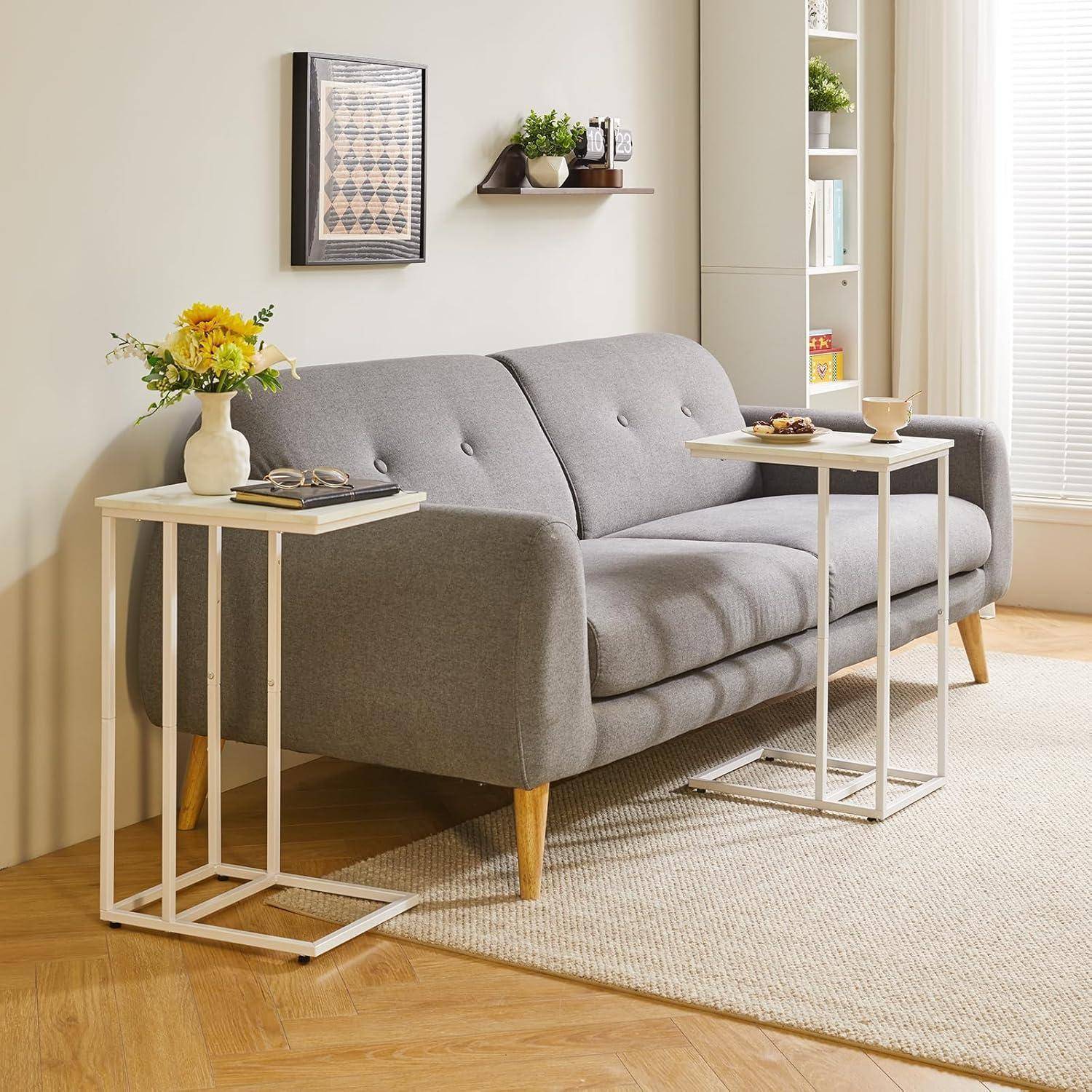 Ein modernes Wohnzimmer mit einem grauen Sofa, zwei Beistelltischen mit Büchern und einer Topfpflanze sowie einer gelben Blumenvase. Neutrale Dekorfarbtöne.