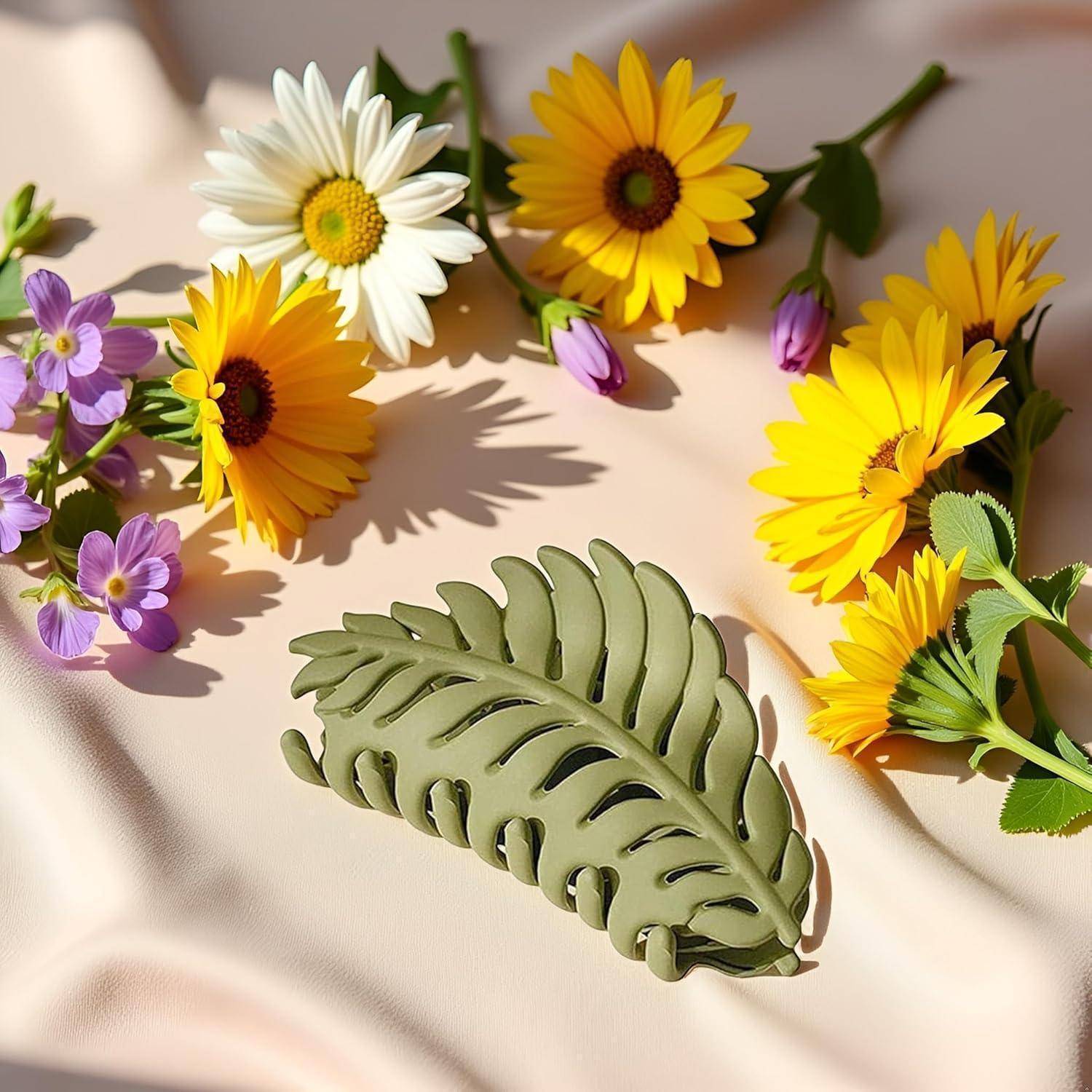 Eine hellgrüne, blattförmige Haarspange liegt auf weichem, cremefarbenem Stoff, umgeben von gelben Gänseblümchen, weißen Gänseblümchen und kleinen lila Blüten.
