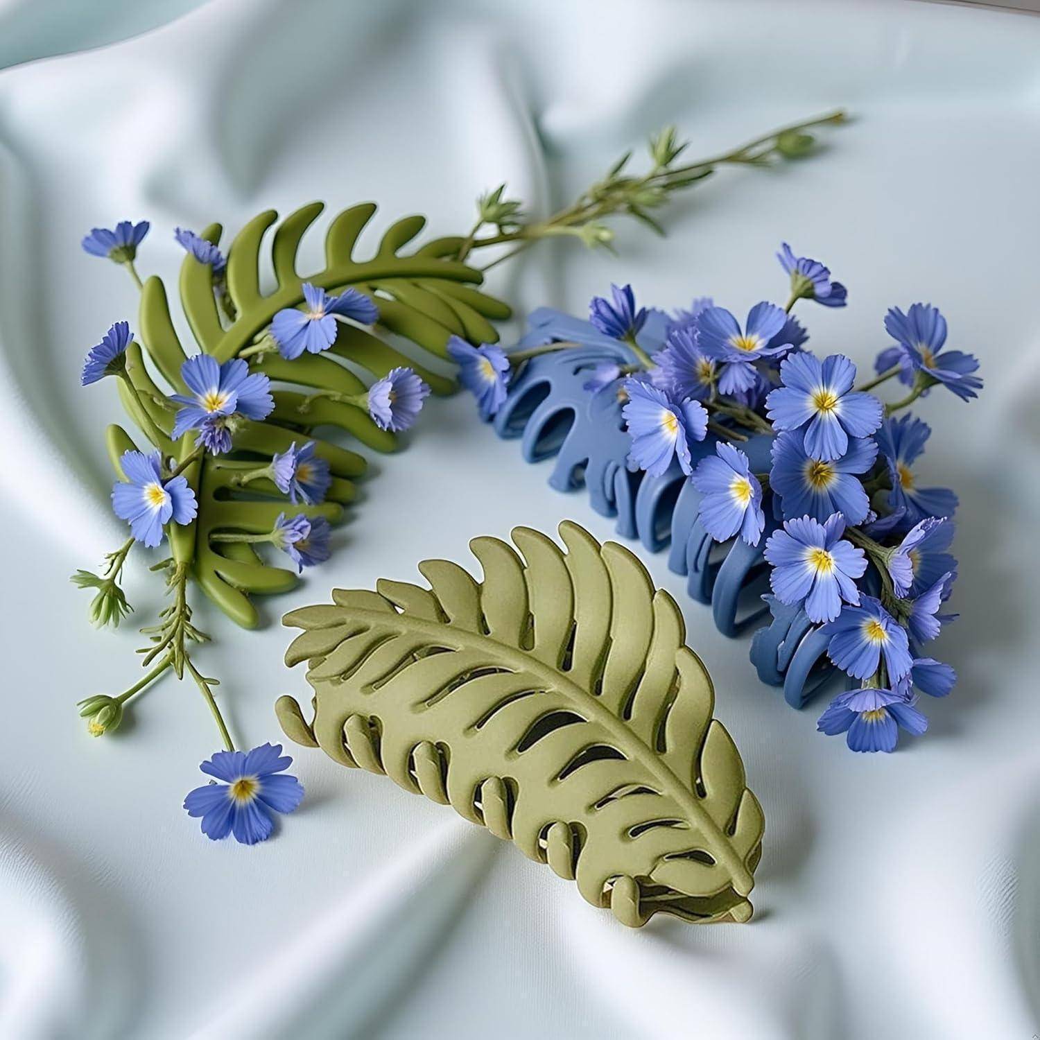 Zwei grüne, blattförmige Haarspangen sind mit kleinen blauen Blumen geschmückt und liegen auf einem hellblauen Stoffhintergrund.