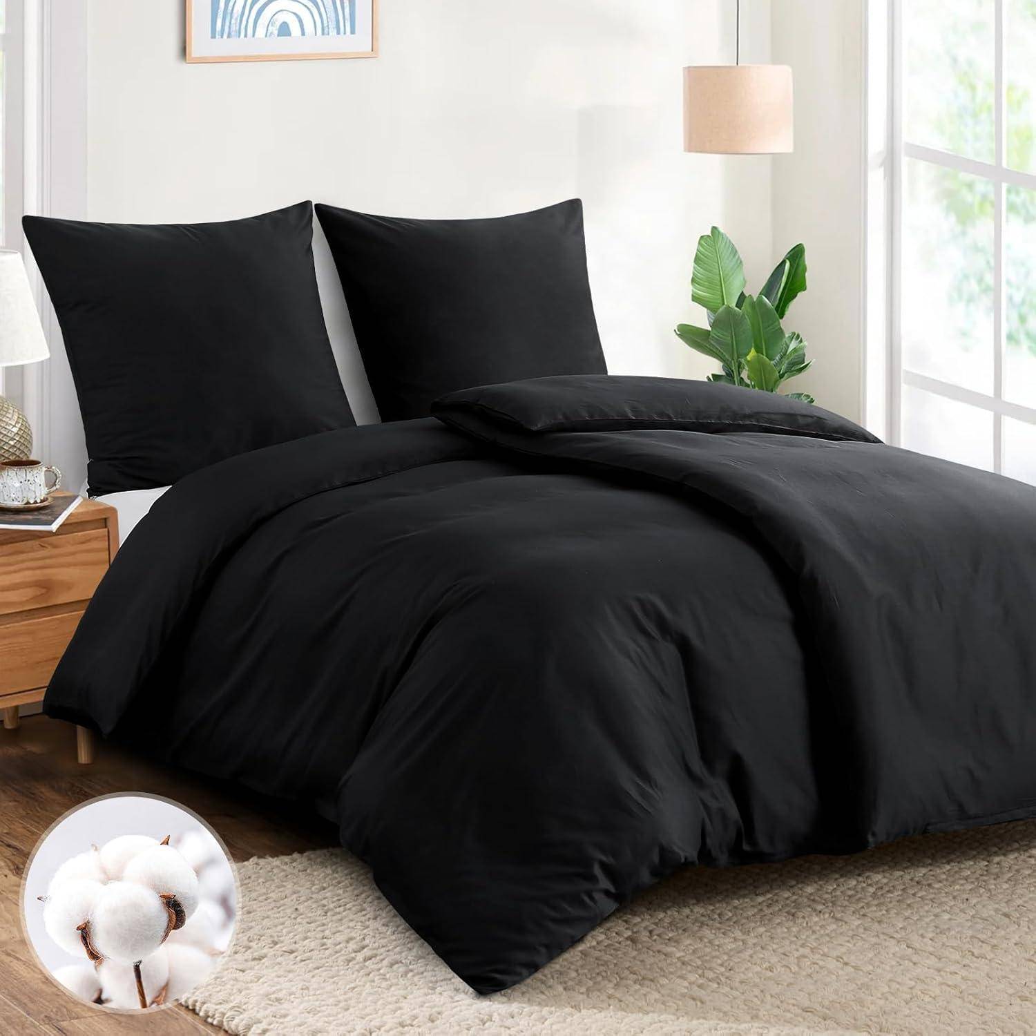 Ein ordentlich gemachtes Bett mit schwarzer Bettwäsche, zwei schwarzen Kissen und einer schwarzen Bettdecke. Eine Lampe steht in der Nähe. Eine Baumwollpflanze ist in einem kleinen Bild zu sehen.