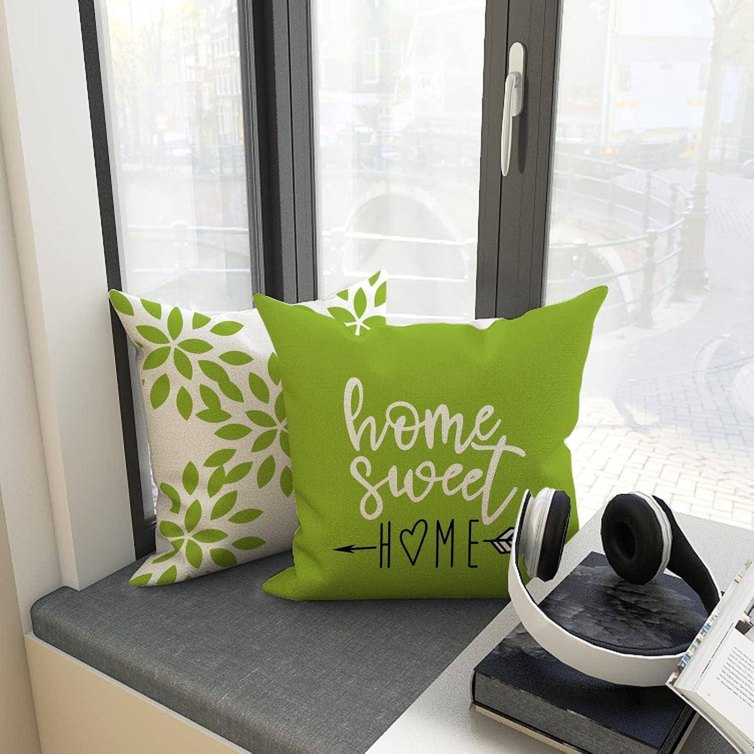 Zwei Kissen auf einer Fensterbank; eines grün mit der Aufschrift ‚home sweet home'. Kopfhörer liegen in der Nähe auf einem Buch. Sonnige Fensteransicht.