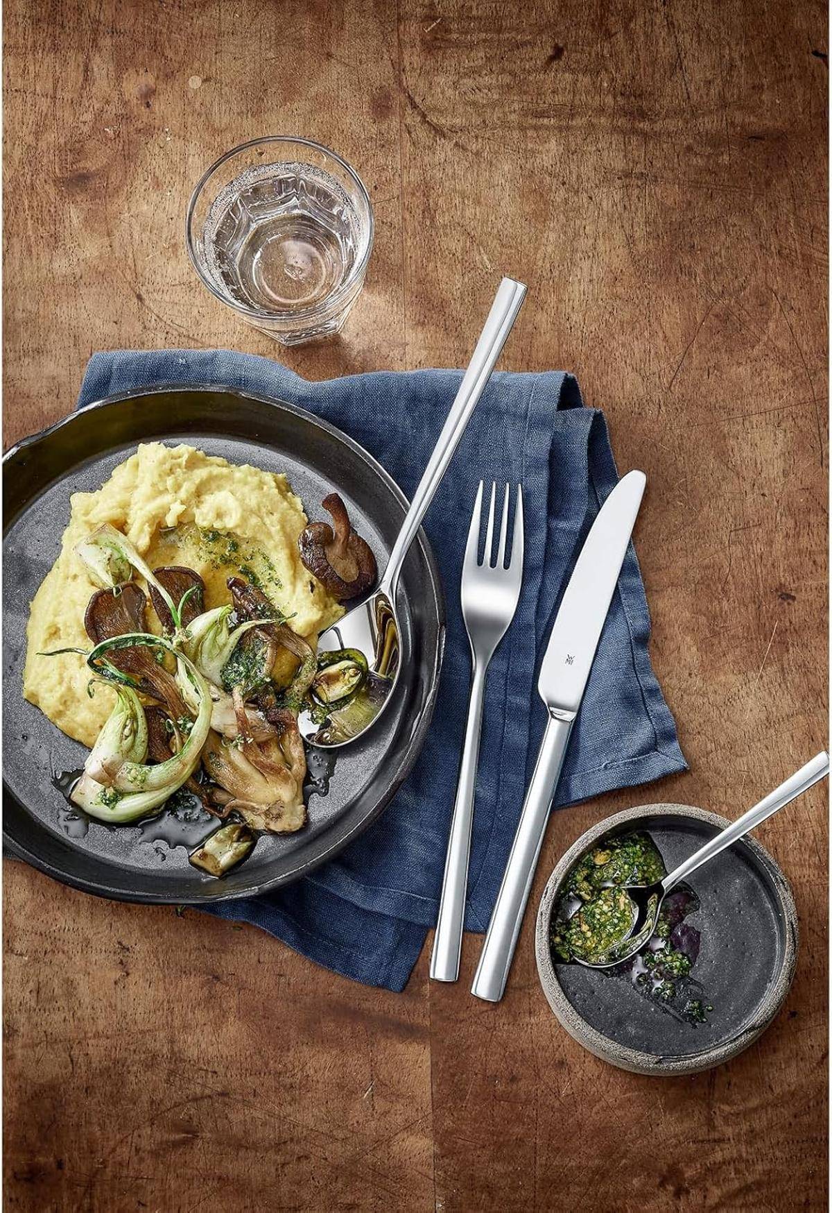 Ein Teller mit Kartoffelpüree, belegt mit gebratenen Gemüsesorten und Kräutern, daneben eine blaue Serviette, Gabel und Messer auf einem Holztisch.