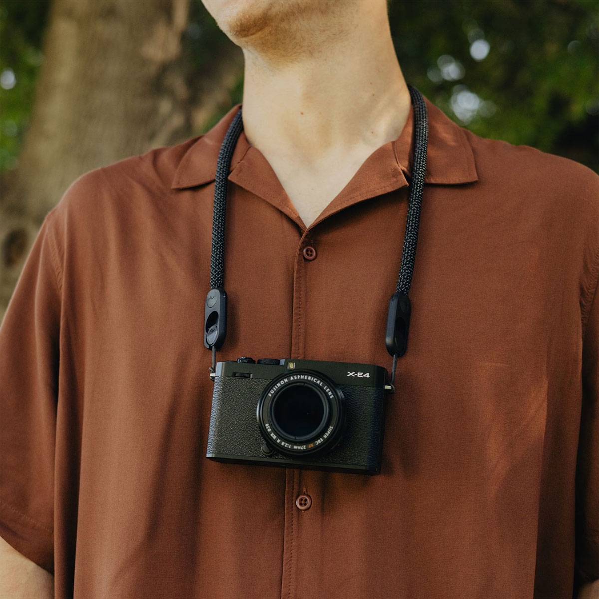 Eine Person mit einem braunen Hemd steht im Freien und trägt eine Kamera um den Hals. Der Hintergrund ist von Grün bewachsen.