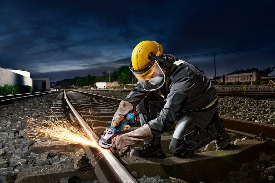 Ein Arbeiter in Schutzausrüstung verwendet ein Schleifwerkzeug auf einer Eisenbahnschiene, umgeben von Schienen und Steinen, mit einem bewölkten Himmel darüber.