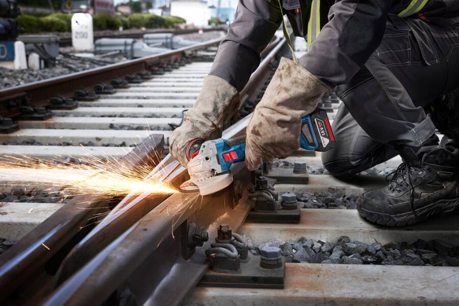 Eine Person verwendet eine Handwinkelschleifer, um eine Eisenbahnschiene zu glätten, wobei Funken sprühen. Die Szene spielt in einem Freiluft-Eisenbahnwartungsbereich.