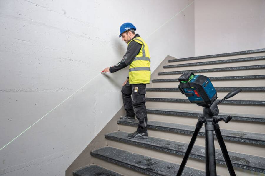 Bauarbeiter verwendet Laser-Nivelliergerät an einer Treppe und stellt Genauigkeit sicher. Er trägt Sicherheitsausrüstung: blauen Helm, gelbe Warnweste. Ein Stativ steht in der Nähe.