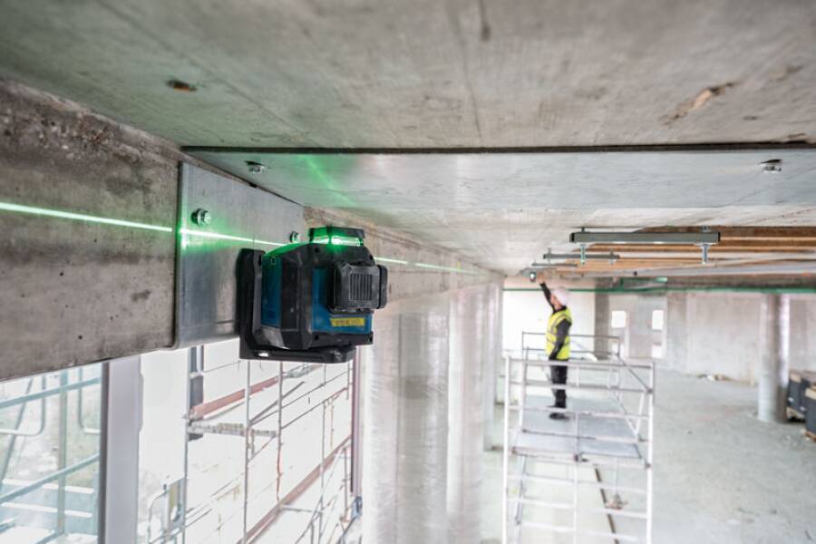 Ein Lasernivelliergerät projiziert eine grüne Linie auf eine Betondecke während der Bauausführung, mit einem Arbeiter im Hintergrund auf einem Gerüst.