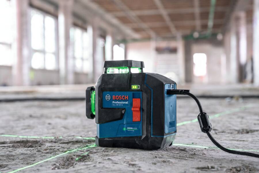 Ein Bosch-Lasernivelliergerät projiziert grüne Linien auf einen Betonboden in einem großen, unfertig wirkenden Raum mit sichtbaren Balken und freigelegten Wänden.