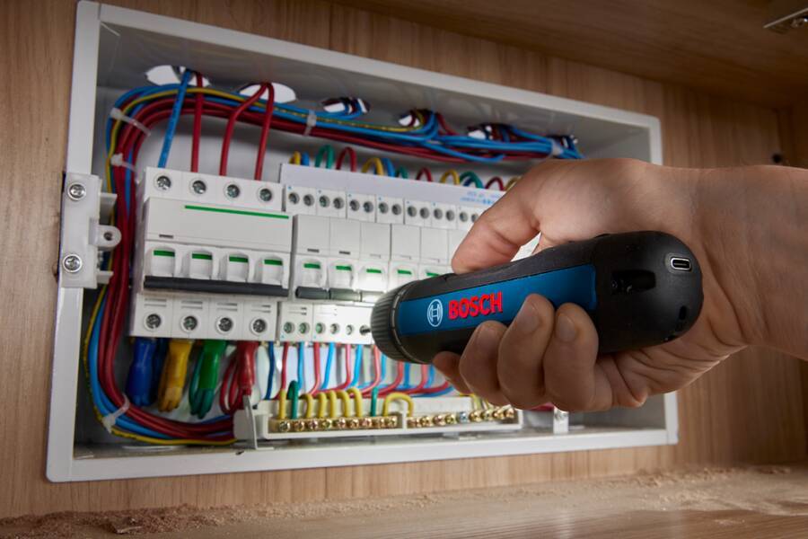 Eine Hand hält einen Bosch-Spannungsprüfer in der Nähe von Sicherungen in einer elektrischen Schalttafel und überprüft die Verbindungen mit farbigen Kabeln.