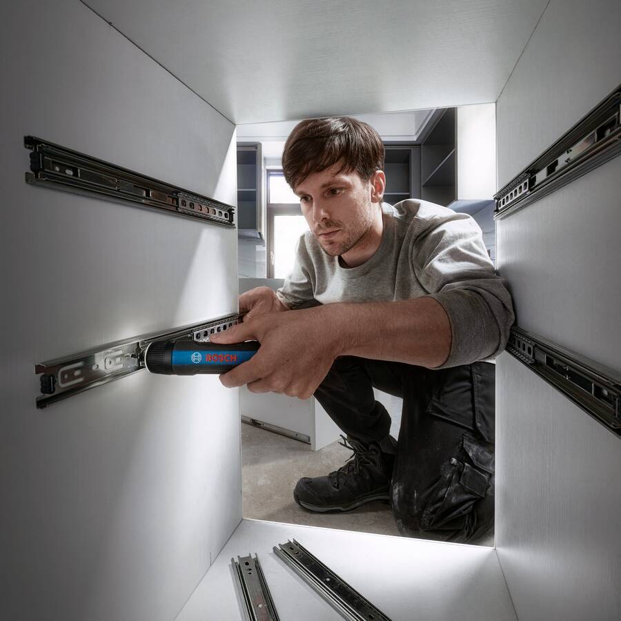 Eine Person installiert eine Schubladenführungsschiene in einem Schrank und verwendet dabei ein blaues Werkzeug. Im Hintergrund sind Regale und andere Schrankteile zu sehen.