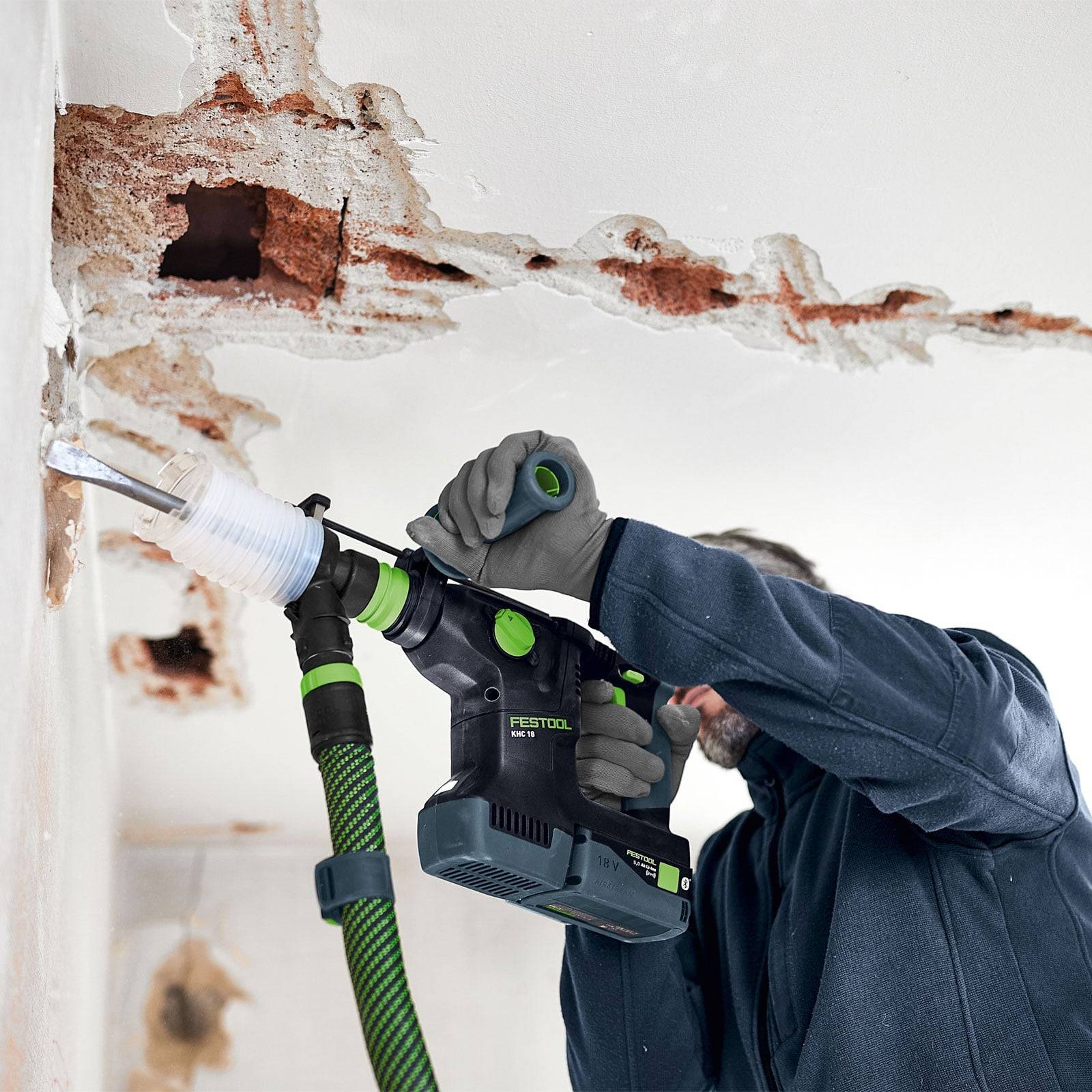 Eine Person, die mit einem Elektrowerkzeug eine beschädigte, abblätternde Wand reinigt oder schleift und dabei Schutzhandschuhe trägt. Die Wand weist deutliche Anzeichen von starkem Verschleiß auf.