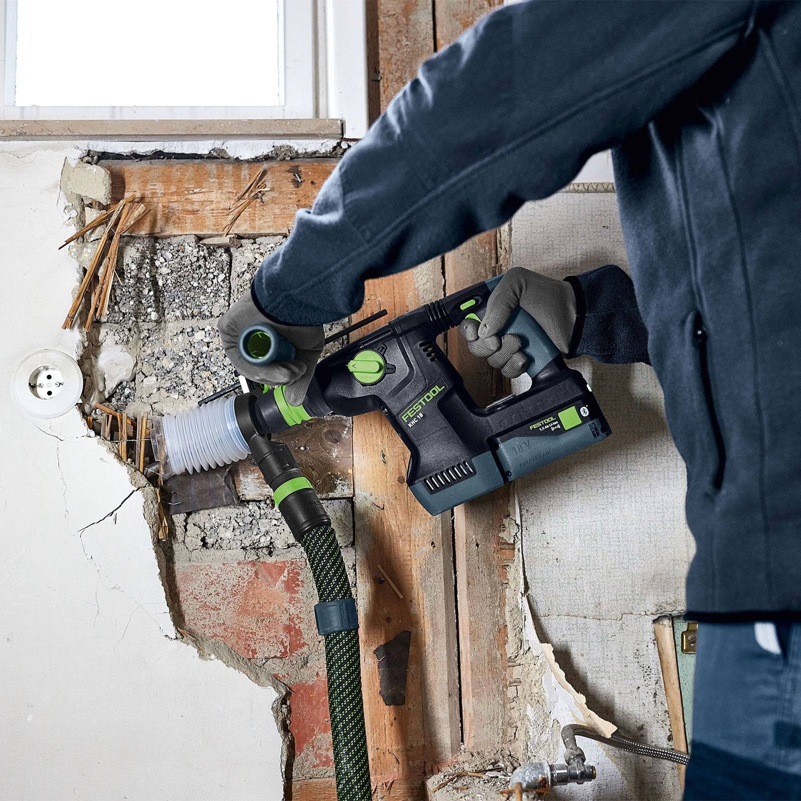 Eine Person in Arbeitskleidung verwendet ein Elektrowerkzeug an einer Wand, die teilweise vom Putz befreit ist und Ziegelsteine und Verkabelung freilegt.