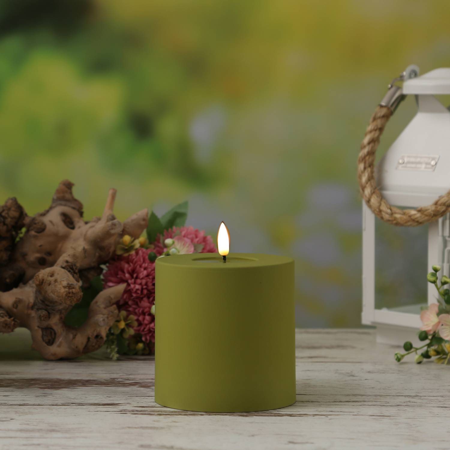 Eine grüne Kerze brennt auf einem Holztisch, umgeben von dekorativen Blumen und einer weißen Laterne in einer Gartenumgebung.
