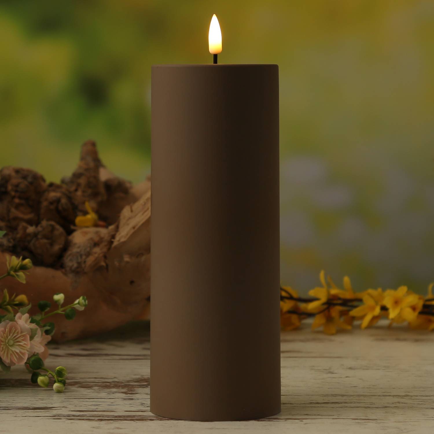 Eine brennende braune Kerze auf einem Holztisch, umgeben von kleinen Blumen und einem verschwommenen Hintergrund von Grünpflanzen, die eine friedvolle Atmosphäre schaffen.