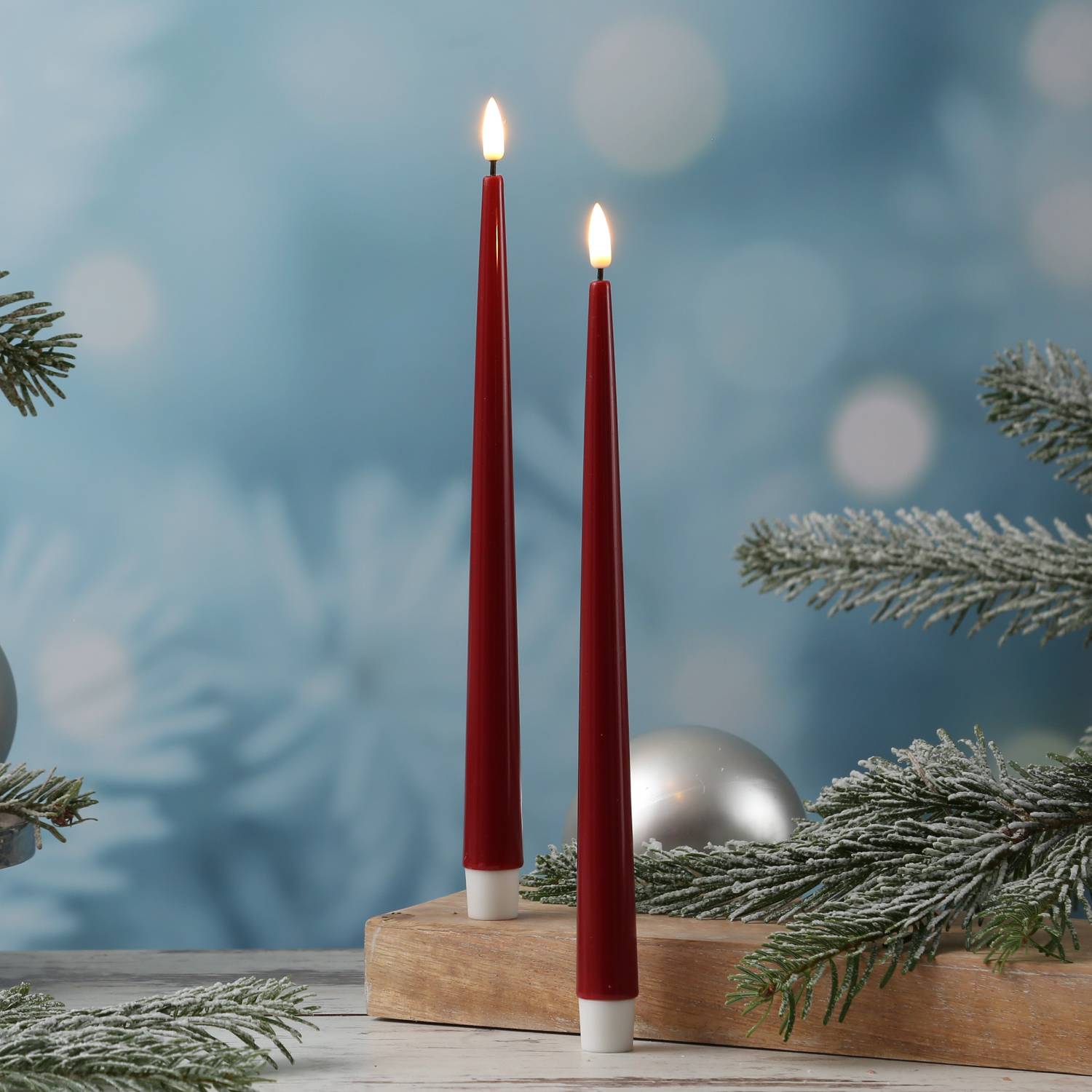 Zwei rote Kerzen auf einem Holzständer mit raureifbedeckten Kiefernzweigen und einem unscharfen blauen Hintergrund, was ein winterliches Thema andeutet.