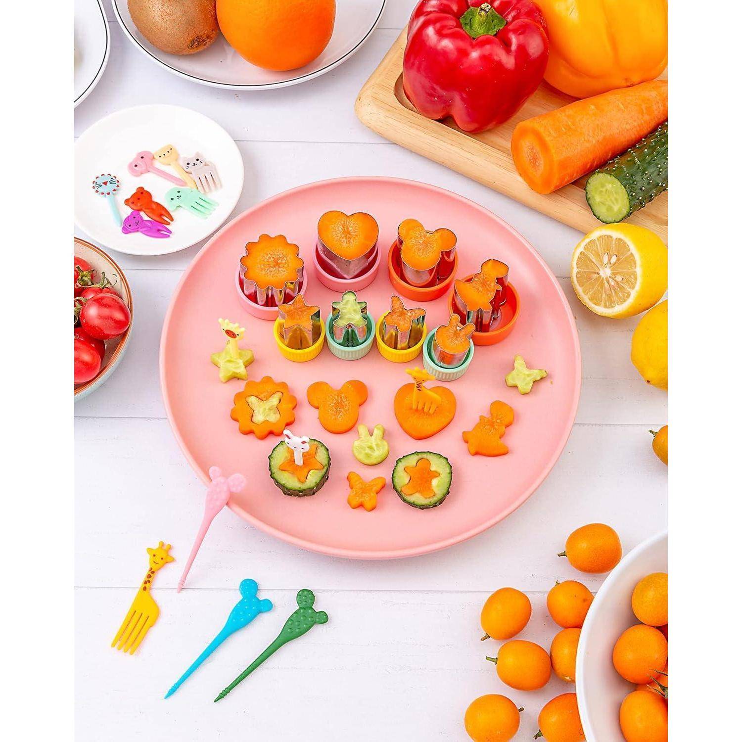Eine Platte mit verschiedenen Obst- und Gemüseformen, darunter Sterne und Tiere, umgeben von Tomaten, Paprika, einer Zitrone und bunten Gabeln.