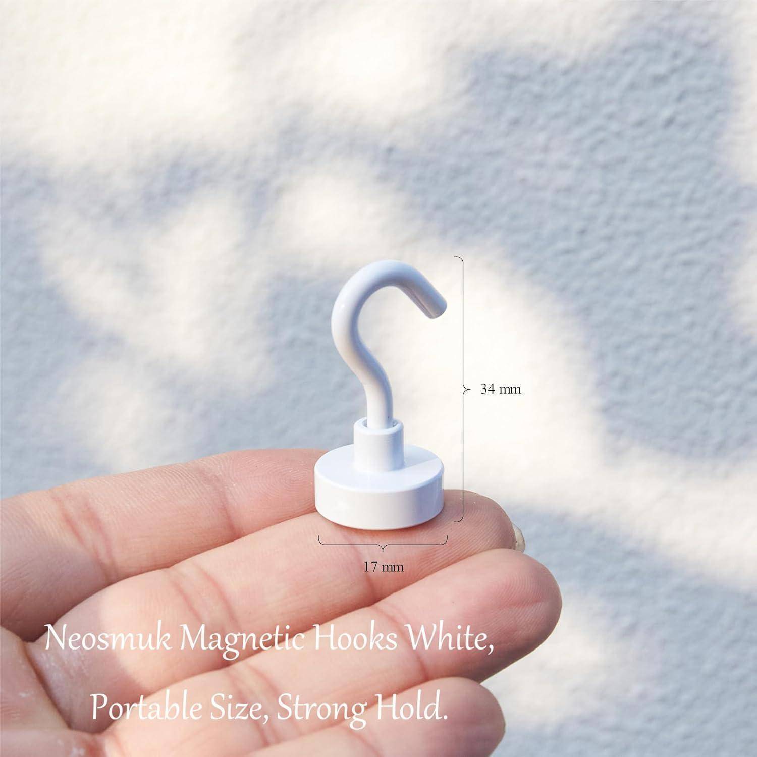 Eine Hand hält einen weißen Neosmuk Magnethafen an einer hellen Wand. Hakendimensionen beschriftet: 34 mm Höhe, 17 mm Basis.