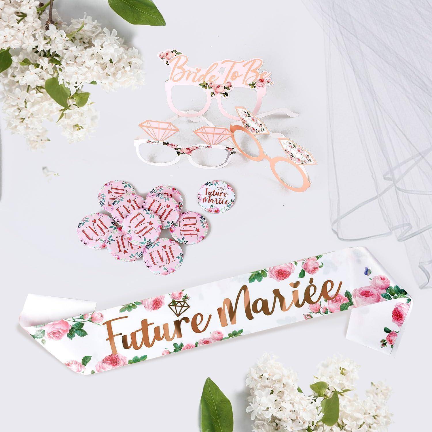 Hochzeitsdekoration auf einer hellen Oberfläche, einschließlich 'Future Mariée' Schärpe, 'Bride to Be' Gläsern, Anstecknadeln und floralen Akzenten.