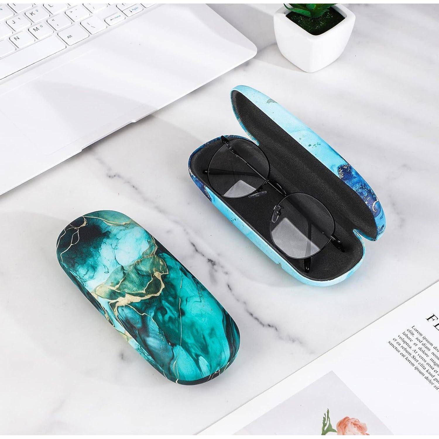 Schwarze Brille in einem marmorierten blau-grünen Etui auf einem weißen Schreibtisch, daneben eine Topfpflanze, ein Laptop und eine Zeitschrift mit einem Blumenbild.