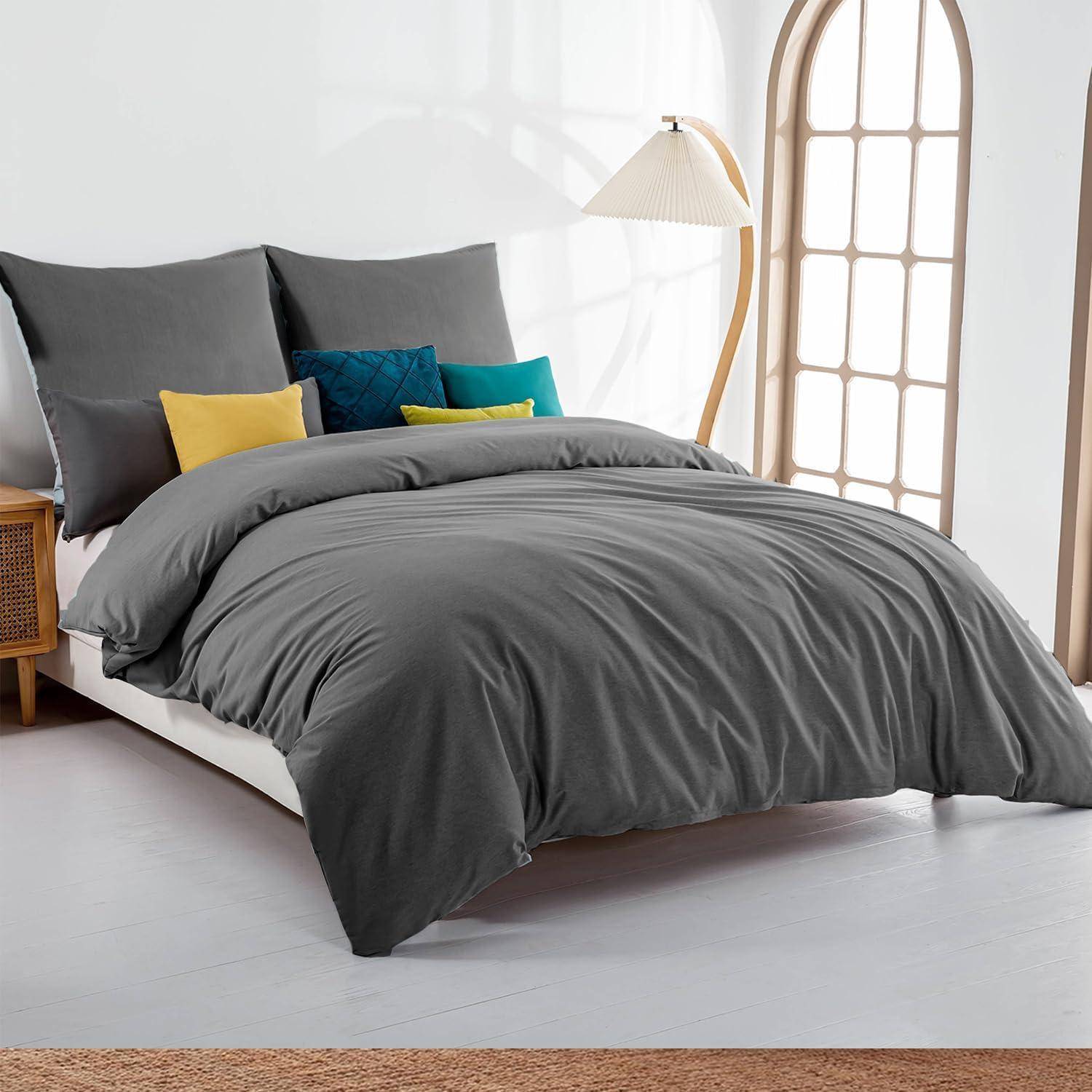 Ein modernes Schlafzimmer mit ordentlich gemachtem Bett, das mit grauer Bettwäsche, dekorativen Kissen, einem Nachttisch aus Flechtwerk und einer hohen Lampe neben einem Bogenfenster ausgestattet ist.
