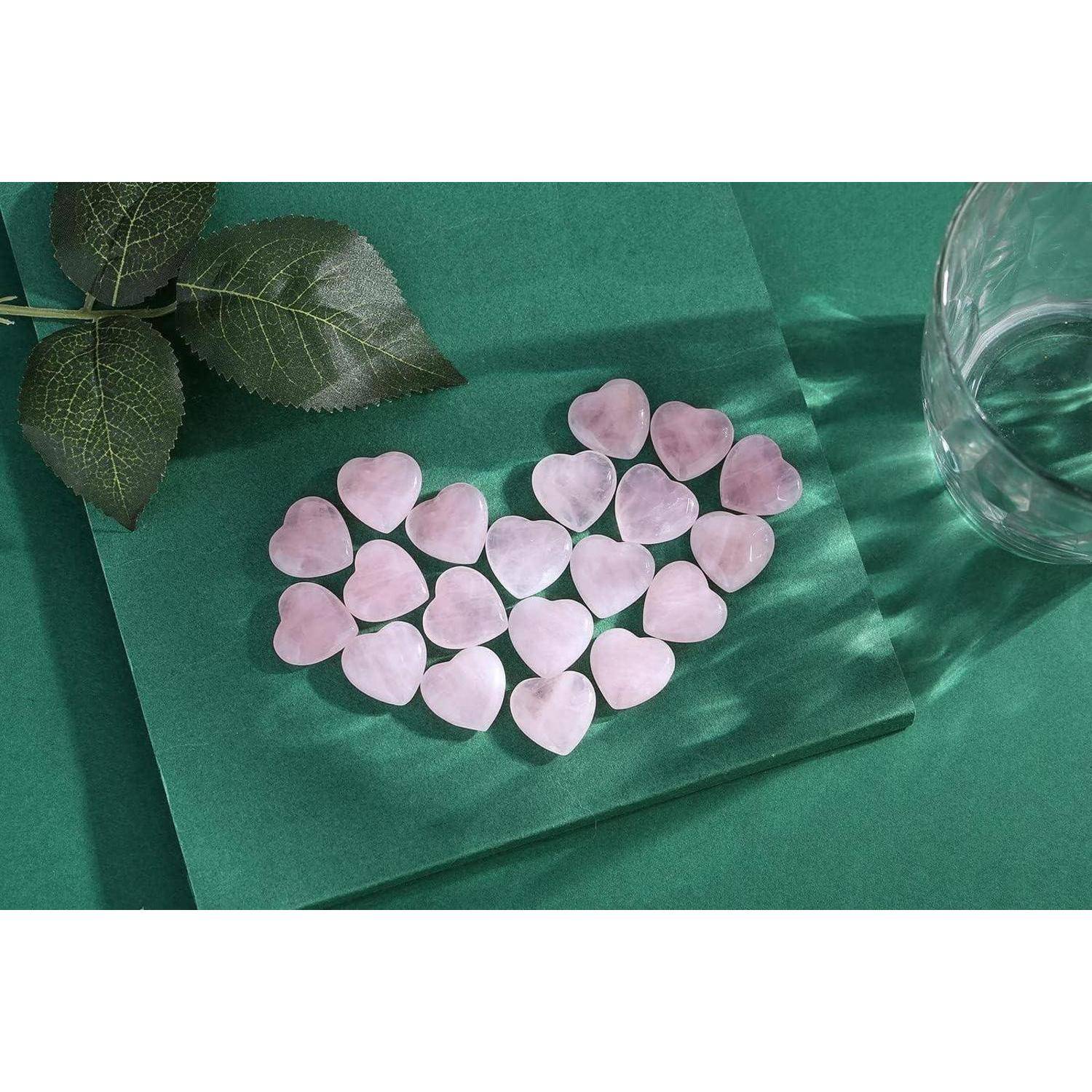Rosenquarz-Herz-Steine sind auf einer grünen Oberfläche angeordnet, neben einem Glas und einem Rosenblatt.
