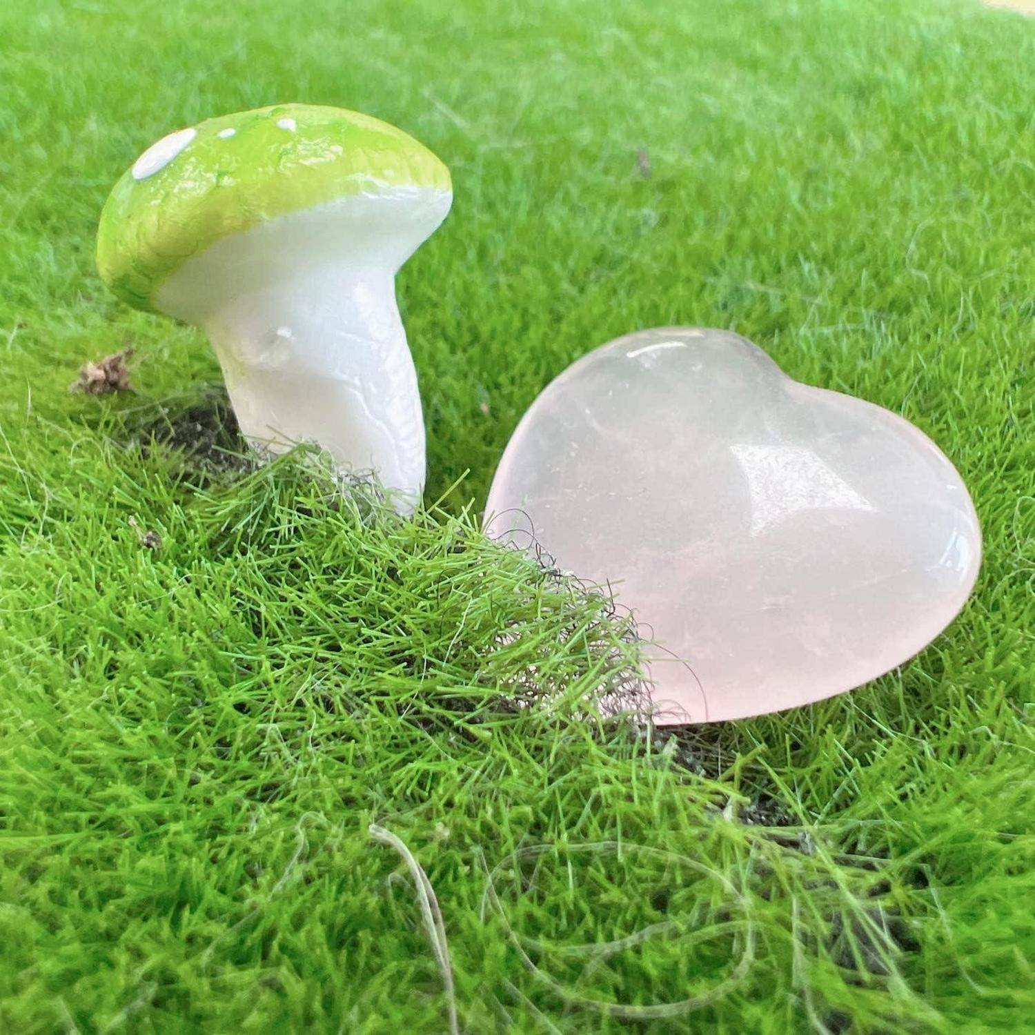 Ein kleiner grüner Pilz und ein rosa herzförmiger Stein ruhen auf hellgrünem Moos und schaffen eine märchenhafte Naturszene.