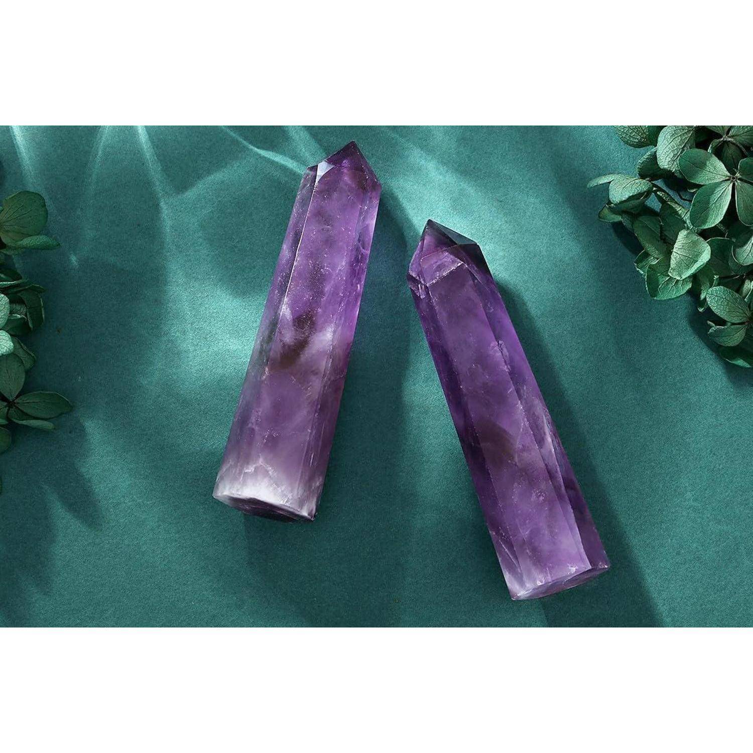 Zwei violette Amethystkristalle auf einer türkisfarbenen Oberfläche, umgeben von grünen Blättern. Das Licht erzeugt Reflexionen auf den Kristallen.