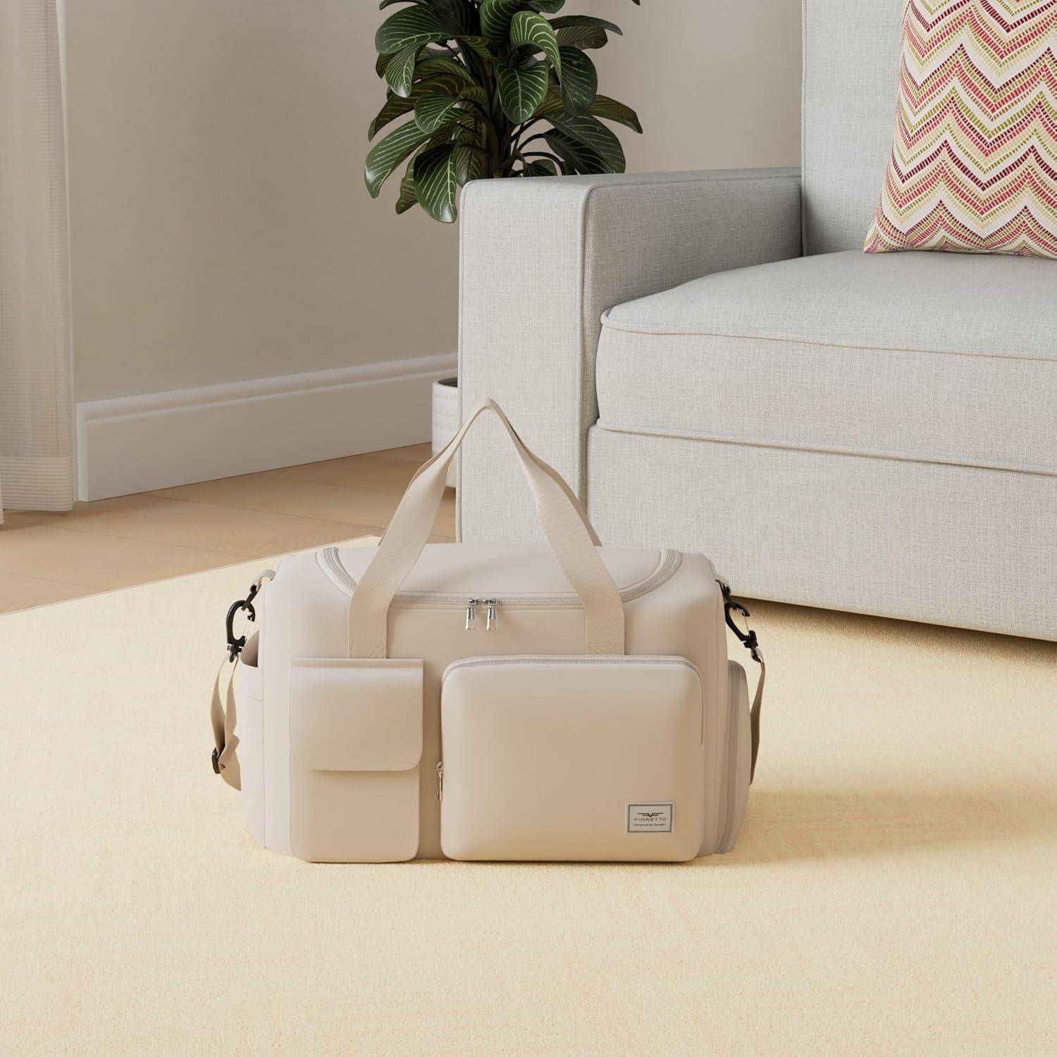 Eine beige Reisetasche mit Taschen steht auf einem hellen Boden neben einem grauen Sofa mit einem Zickzack-gemusterten Kissen in einem lichtdurchfluteten Raum.