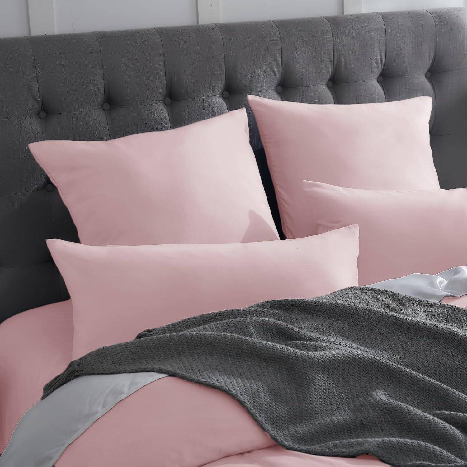 Ein ordentlich gemachtes Bett mit hellrosa Bettwäsche, mehreren rosa Kissen vor einem grauen gepolsterten Kopfteil und einer dunkelgrauen Waffelpikee-Decke.
