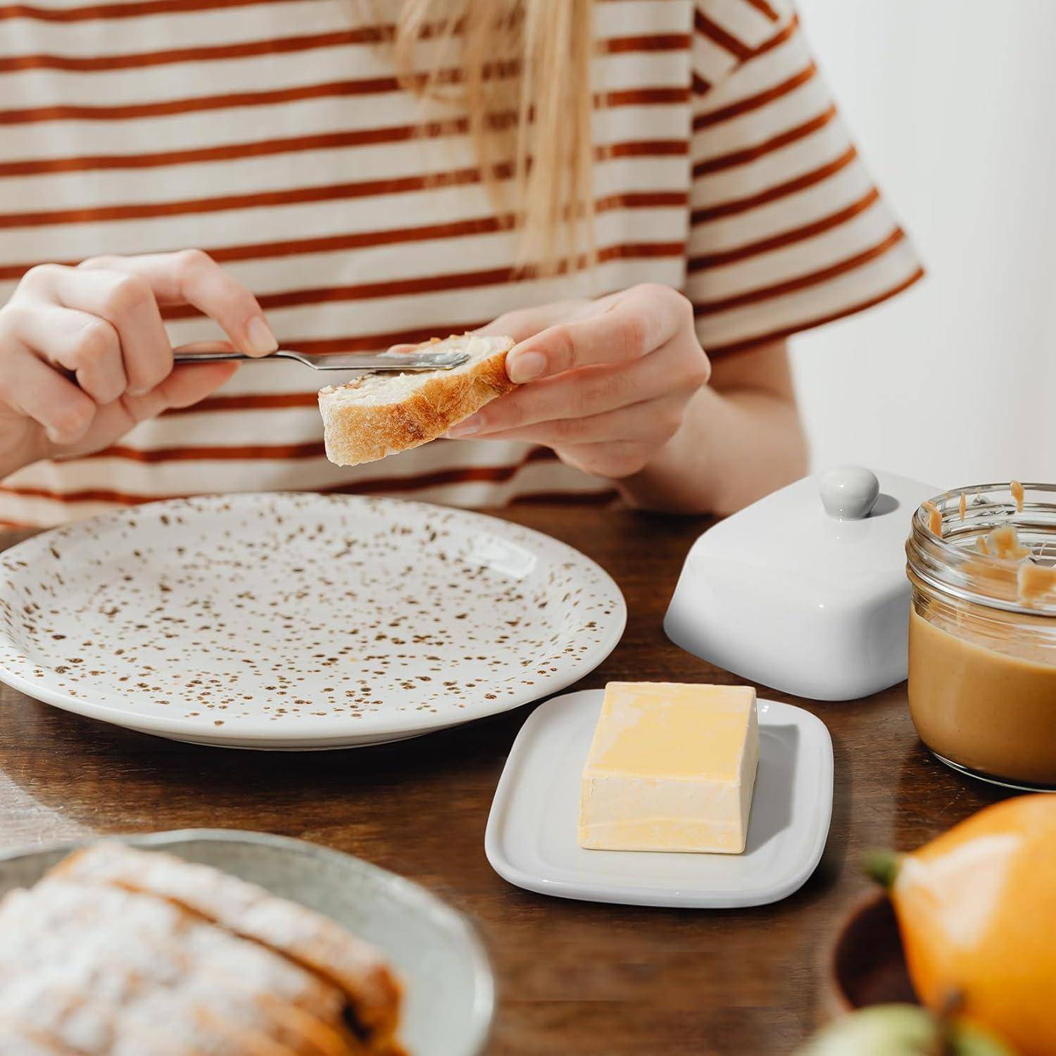 Eine Person streicht Butter auf Toast an einem Frühstückstisch, wobei ein Marmeladenglas, ein Teller mit Gebäck und ein Butterblock sichtbar sind.