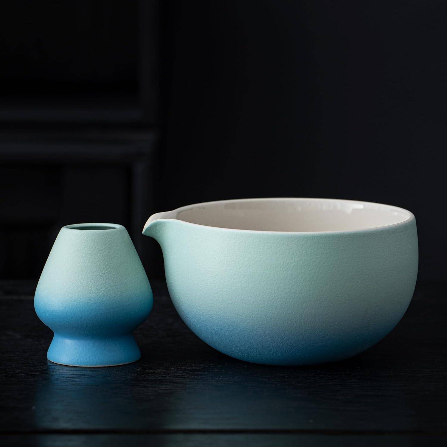 Eine Keramiktasse in Verlaufstönen von Blaugrün und Blau mit einer Ausgussöffnung steht neben einer passenden blauen Vase auf einem dunklen Tisch vor einem dunklen Hintergrund.
