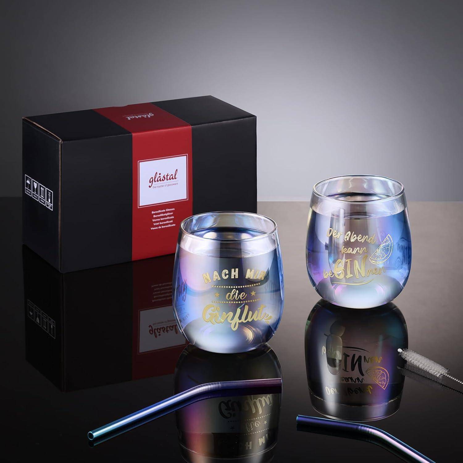 Zwei Glasbecher mit bunten Designs und Text auf Deutsch, neben einer schwarzen und roten Geschenkbox platziert. Ein passendes Trinkhalm liegt davor.