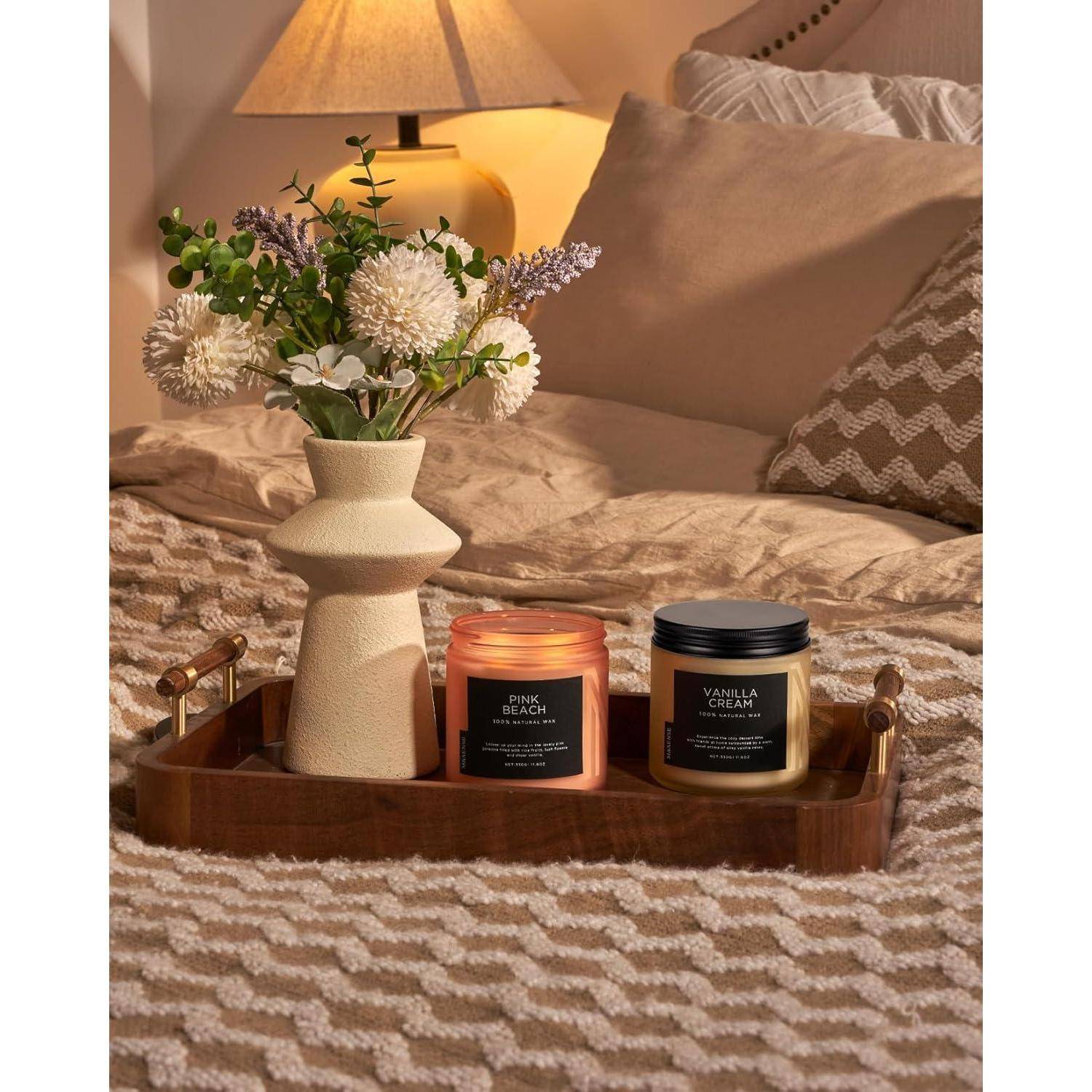 Ein gemütliches Schlafzimmer mit einem Holztablett auf dem Bett, das eine Blumenvase und zwei duftende Kerzen mit den Bezeichnungen „Pink Beach