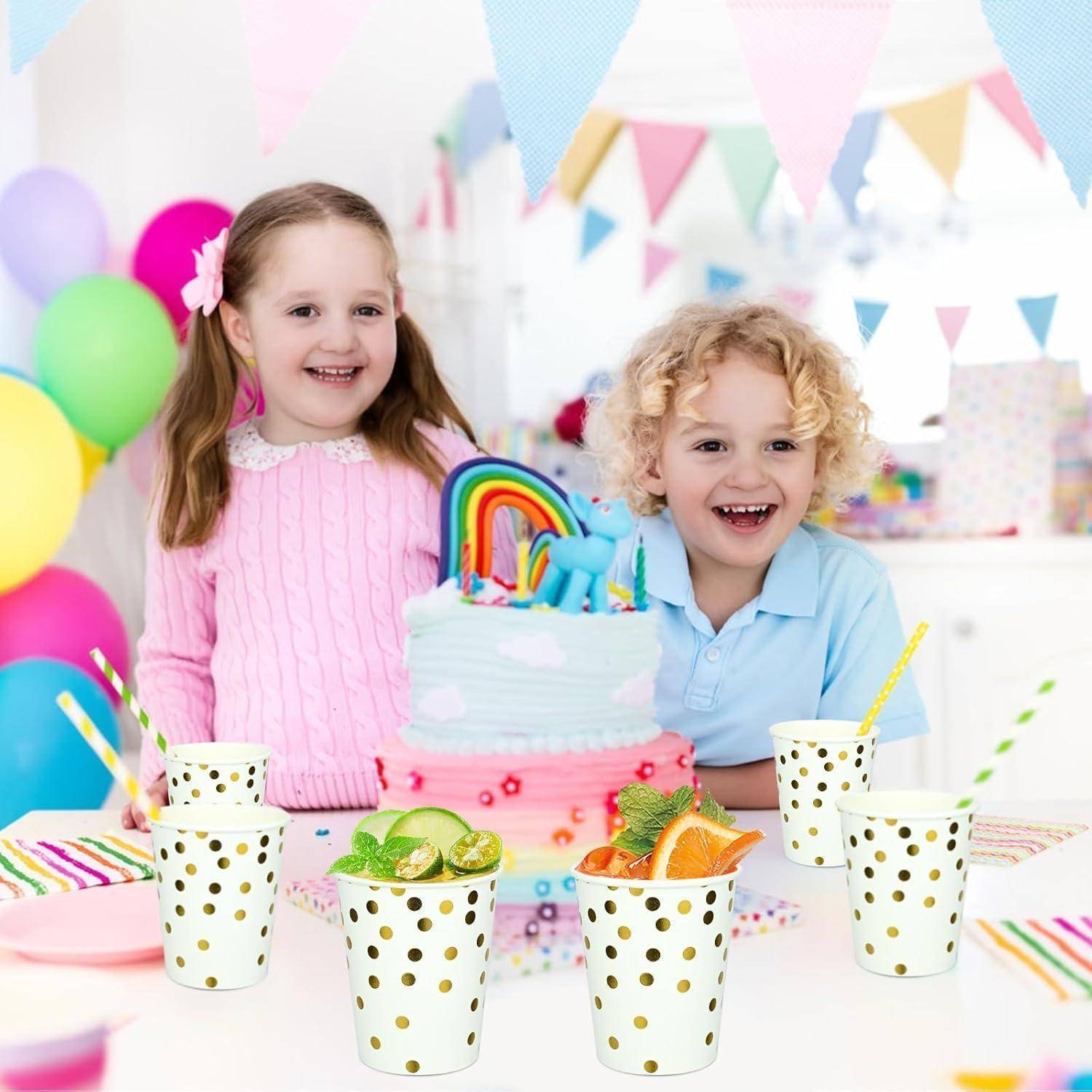 Ein Mädchen und ein Junge feiern Geburtstag in einem festlich geschmückten Raum mit Luftballons. Auf dem Tisch steht eine Torte, die mit einem Regenbogen und Einhörnern verziert ist.