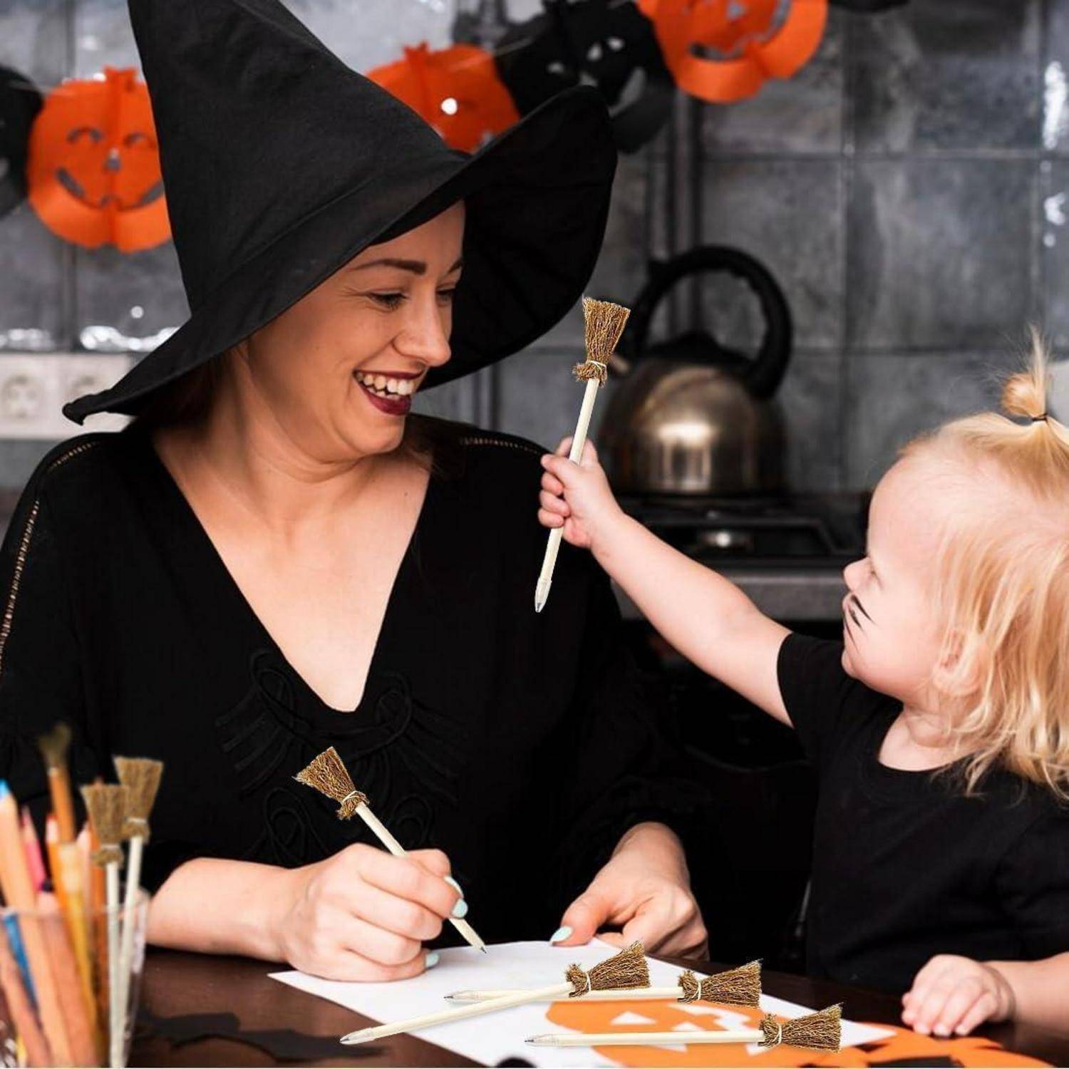 Eine Frau mit einem Hexenhut lächelt, während sie mit einem Kind an einem Tisch zeichnet, der mit Halloween-Bastelarbeiten und Kürbisausschnitten dekoriert ist.