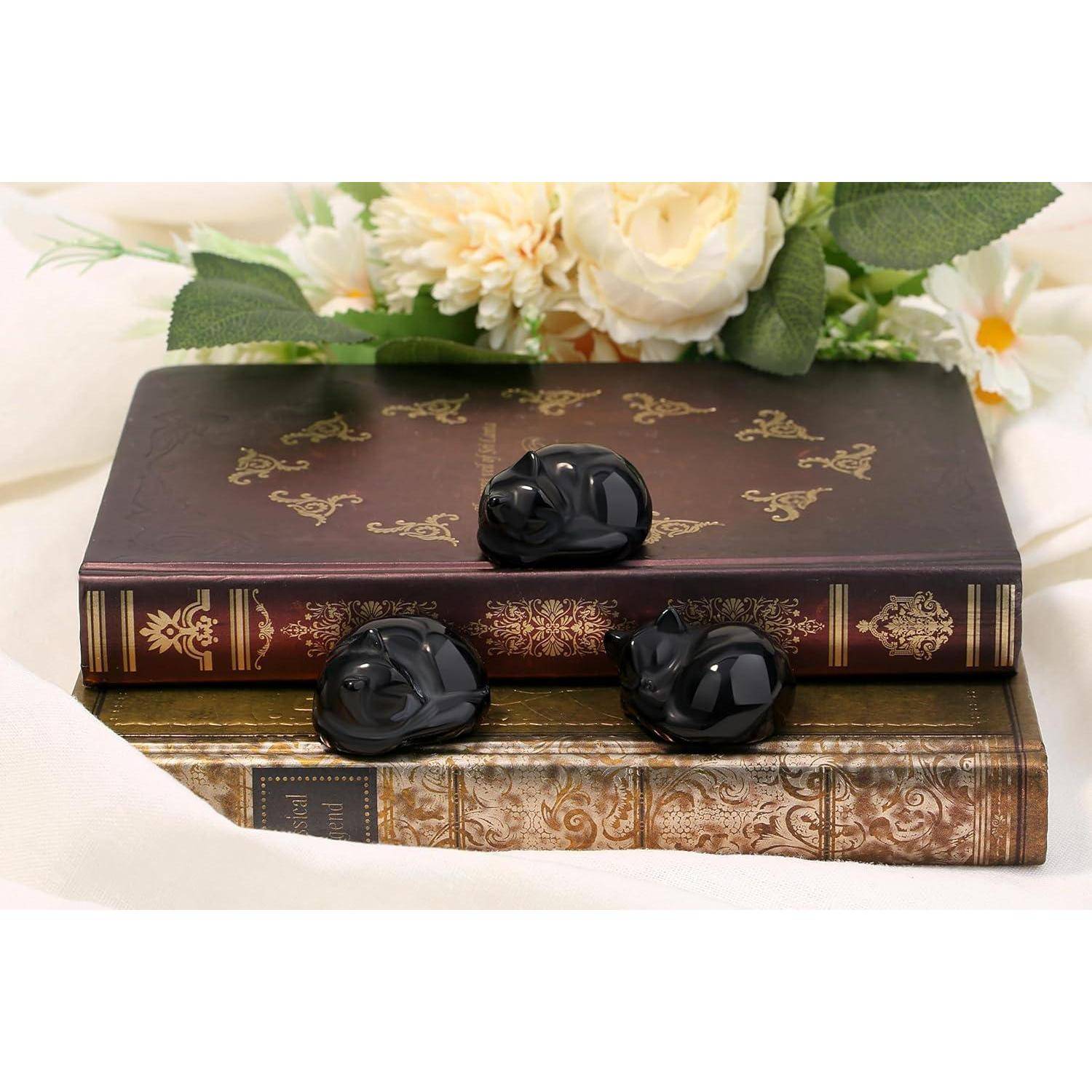 Drei schwarze Katzenfigürchen ruhen auf zwei gestapelten Vintage-Büchern mit kunstvollen Einbänden. Weiße Blumen im Hintergrund verleihen eine dekorative Note.