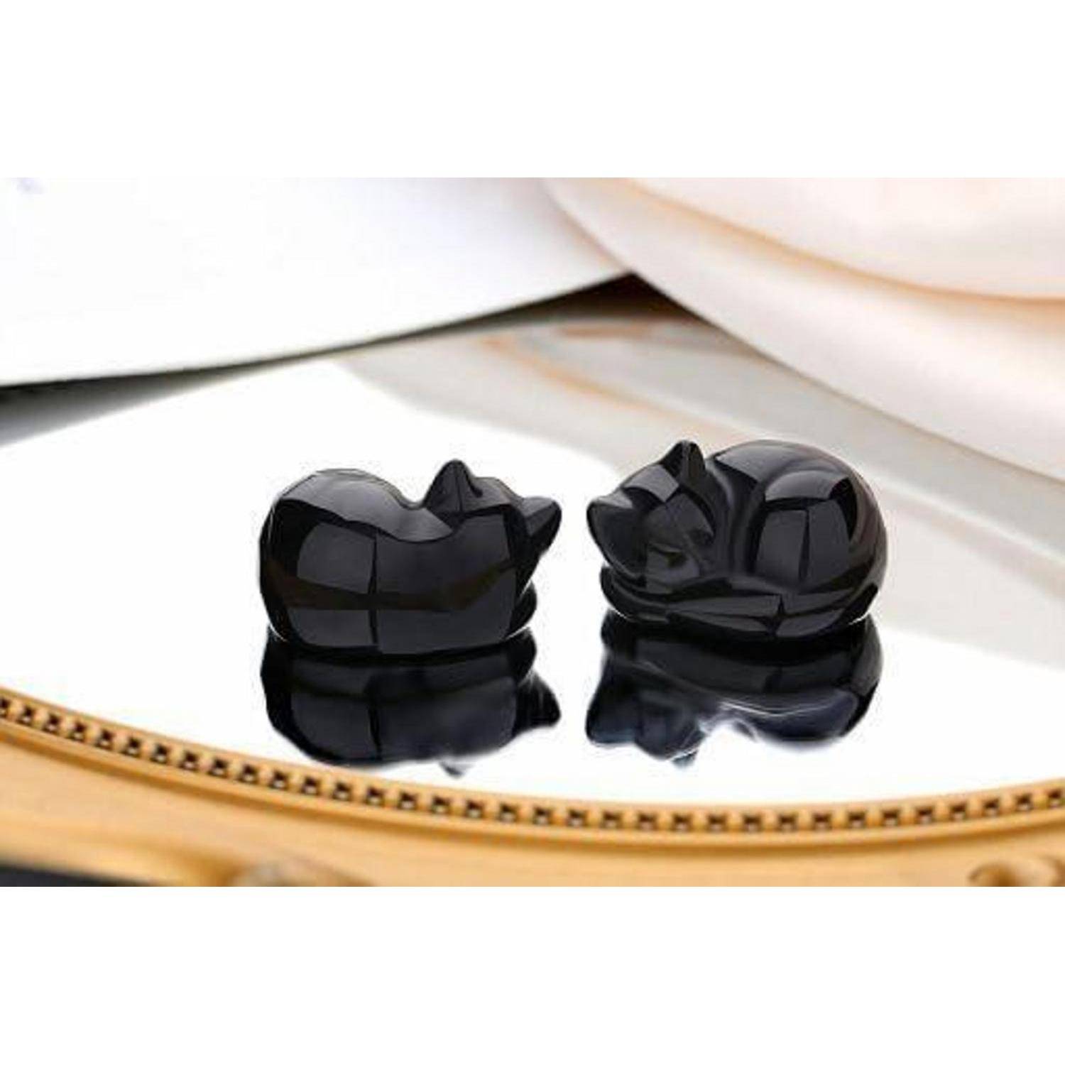 Zwei kleine, glänzend schwarze Katzenfiguren, die auf einer reflektierenden Oberfläche ruhen.