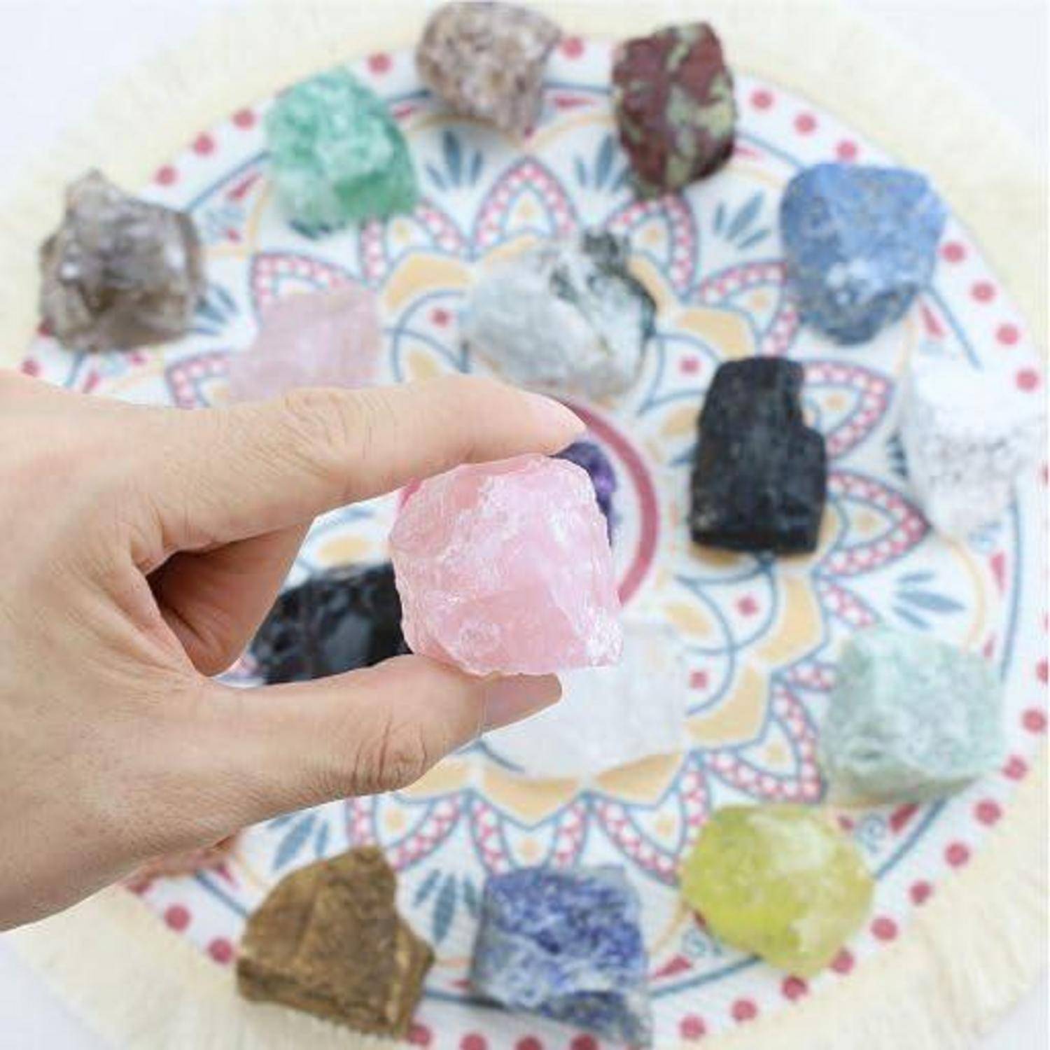 Eine Hand hält einen rosa rauen Edelstein über eine kreisförmige Anzeige mit verschiedenen farbenfrohen, rohen Edelsteinen, die radial auf einem gemusterten Hintergrund angeordnet sind.