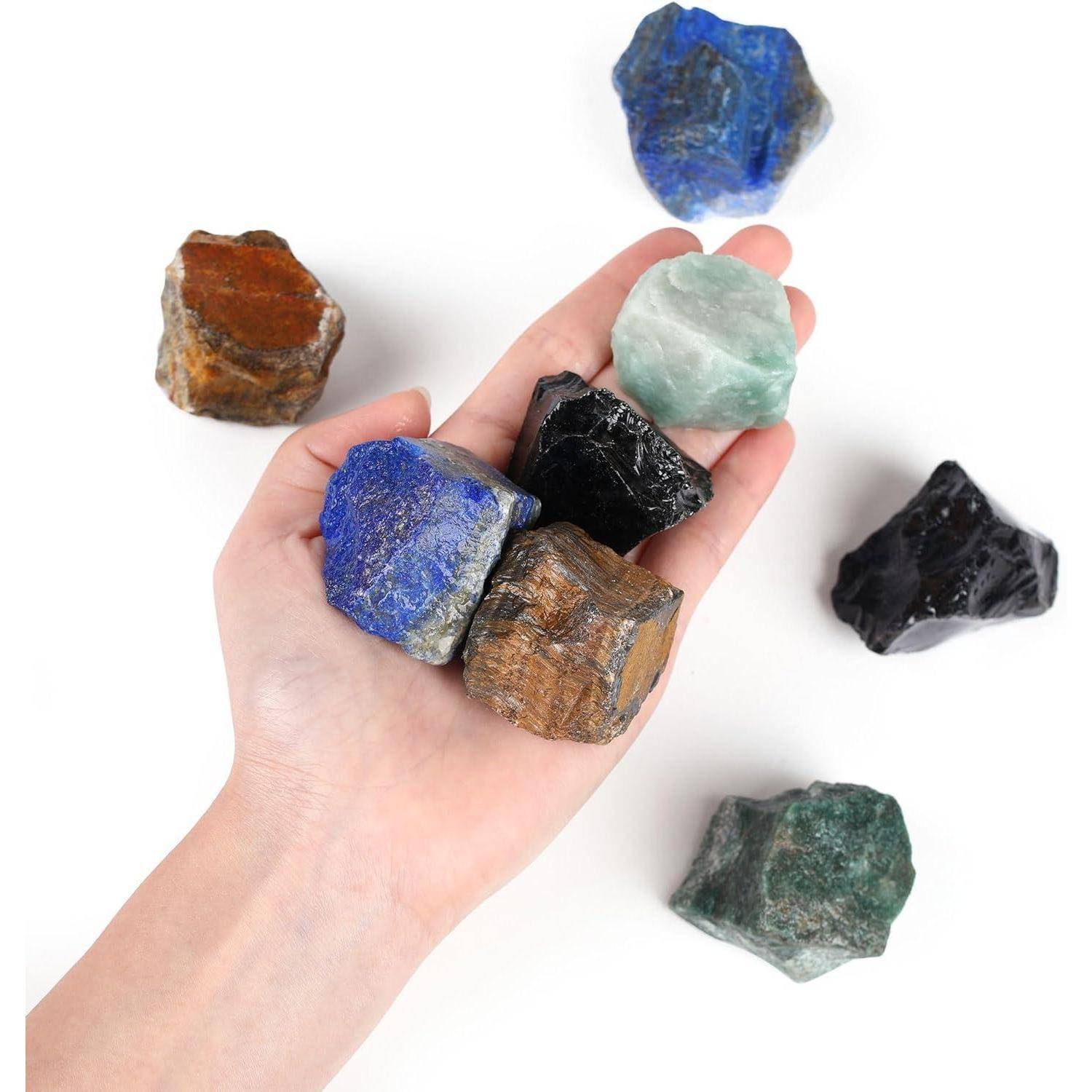 Eine Hand, die verschiedene bunte Rohedelsteine, einschließlich blauer, grüner und brauner Steine, vor einem weißen Hintergrund hält und dabei ihre Texturen betont.