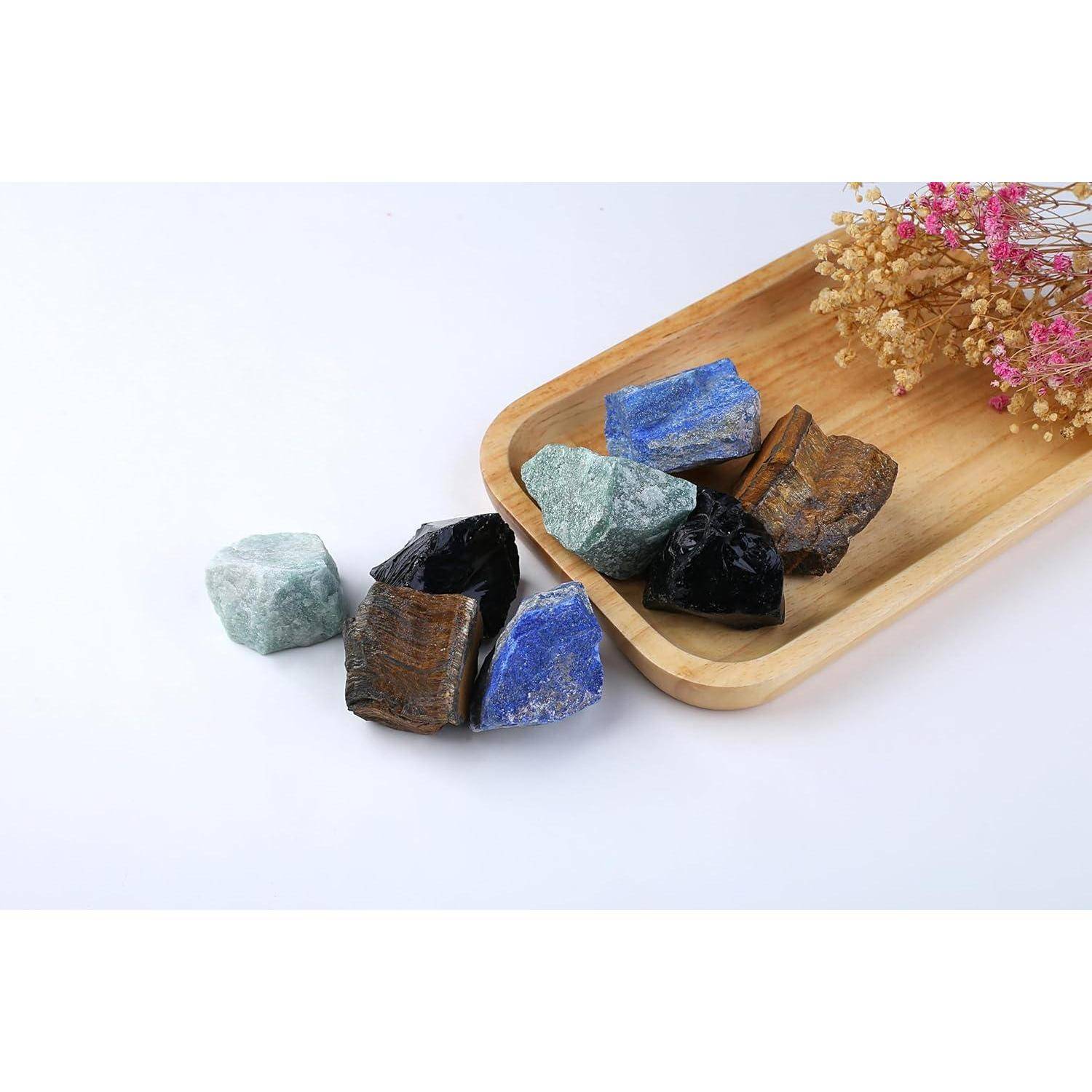 Ein Holztablett mit verschiedenen rauen Steinen in den Farben Blau, Schwarz, Grün und Braun, daneben dekorative getrocknete Blumen auf weißem Hintergrund.