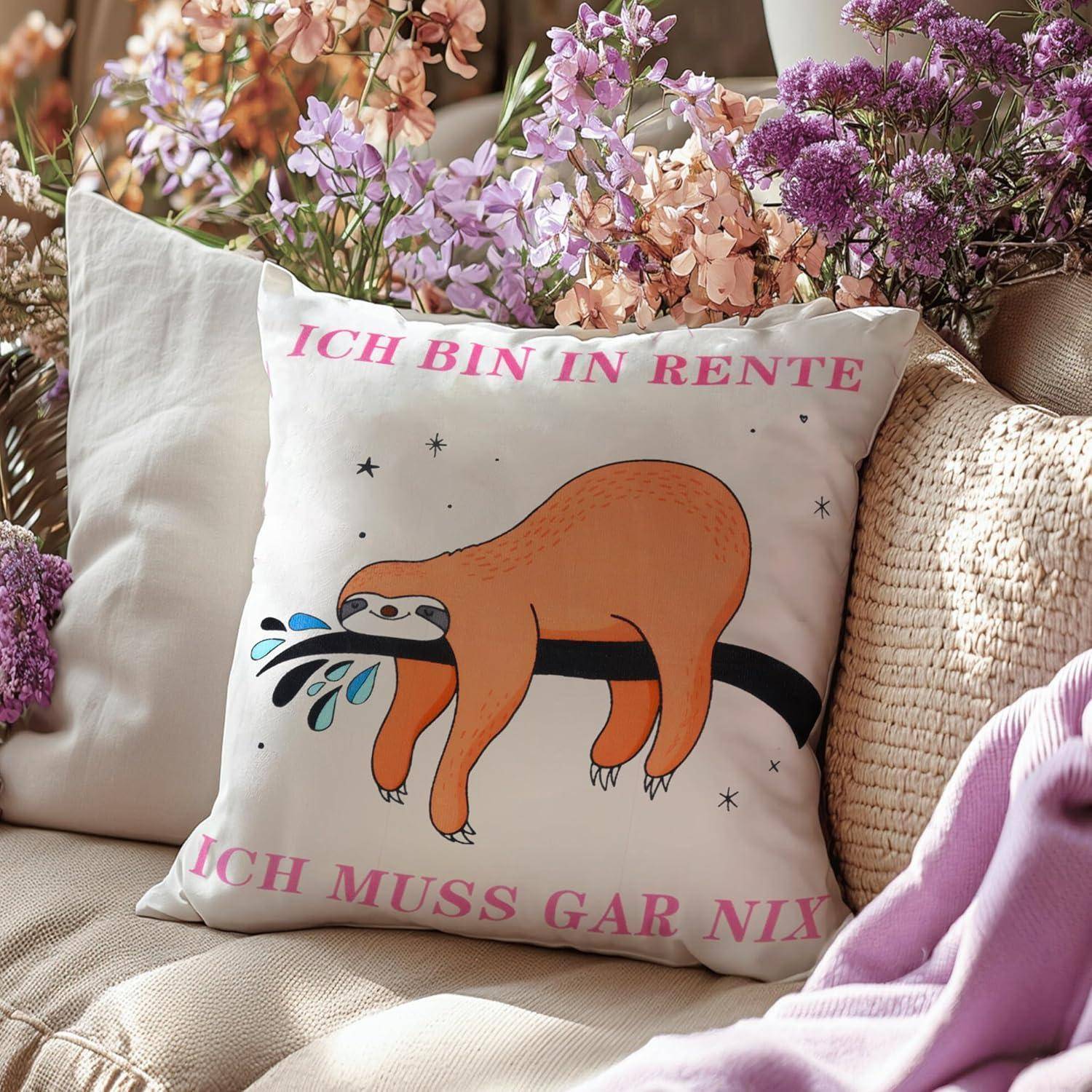 Ein Kissen auf einem Sofa zeigt einen Cartoon-Faultier, der an einem Ast hängt, mit dem Text ‚Ich bin in Rente, ich muss gar nix', umgeben von Blumen.