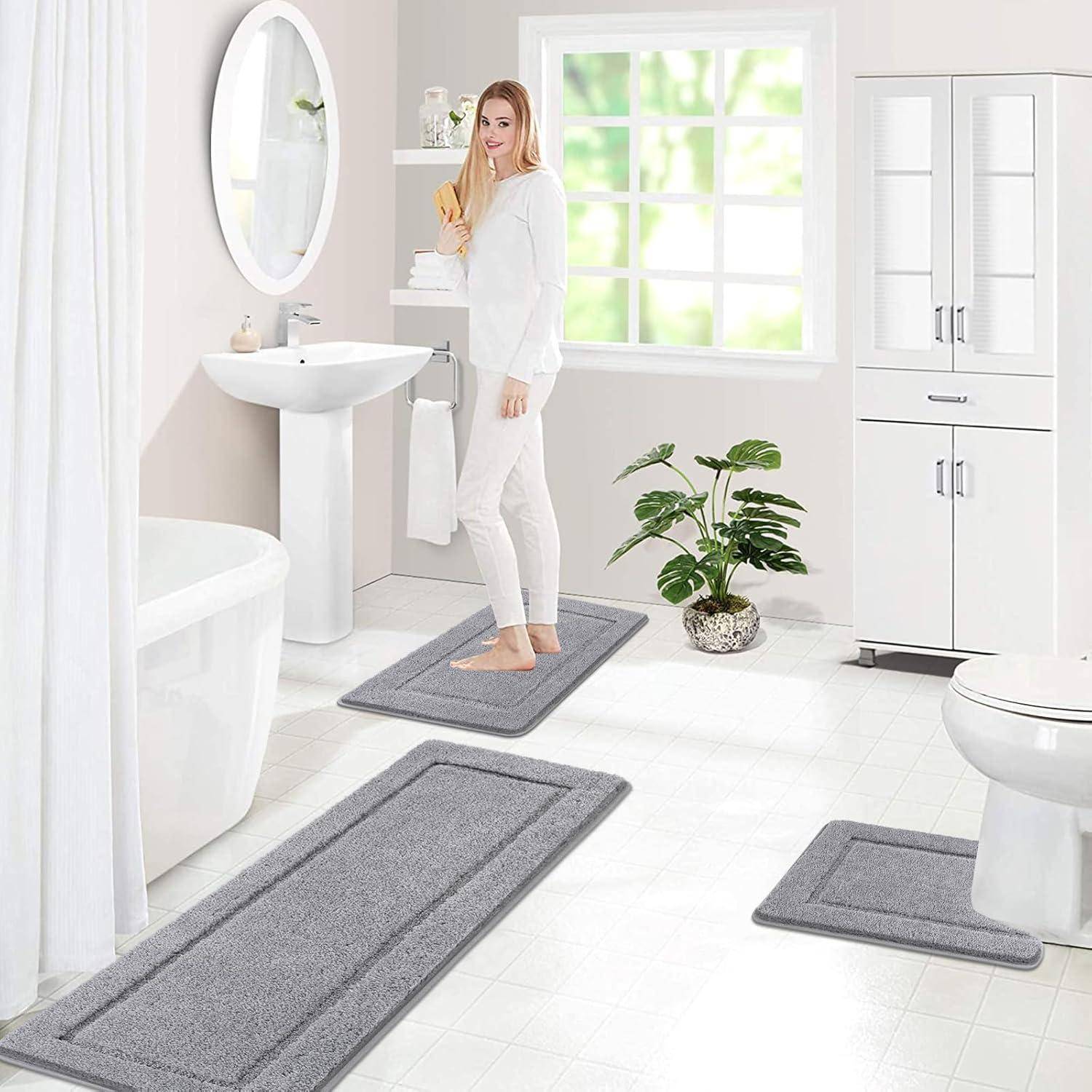 Eine Frau in einem weißen Badezimmer steht auf einem grauen Teppich in der Nähe eines Waschbeckens, wobei eine Badewanne, eine Toilette und eine Topfpflanze zu sehen sind. Sonnenlicht filtert durch ein Fenster.