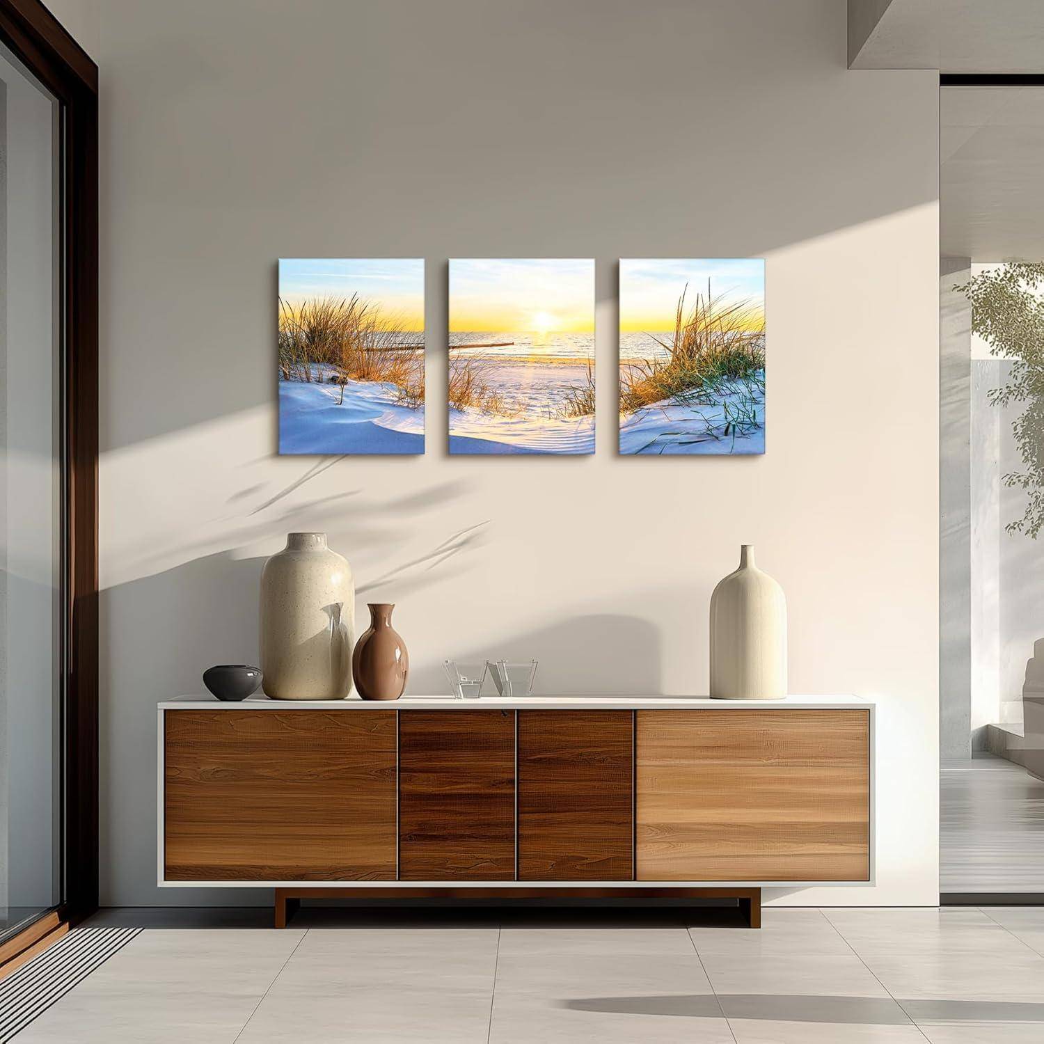 Ein moderner Raum zeigt ein schlankes Holzsideboard, geschmückt mit zwei Vasen. Ein dreiteiliges Kunstwerk darüber stellt eine friedvolle Strand-Sonnenuntergangsszene dar.