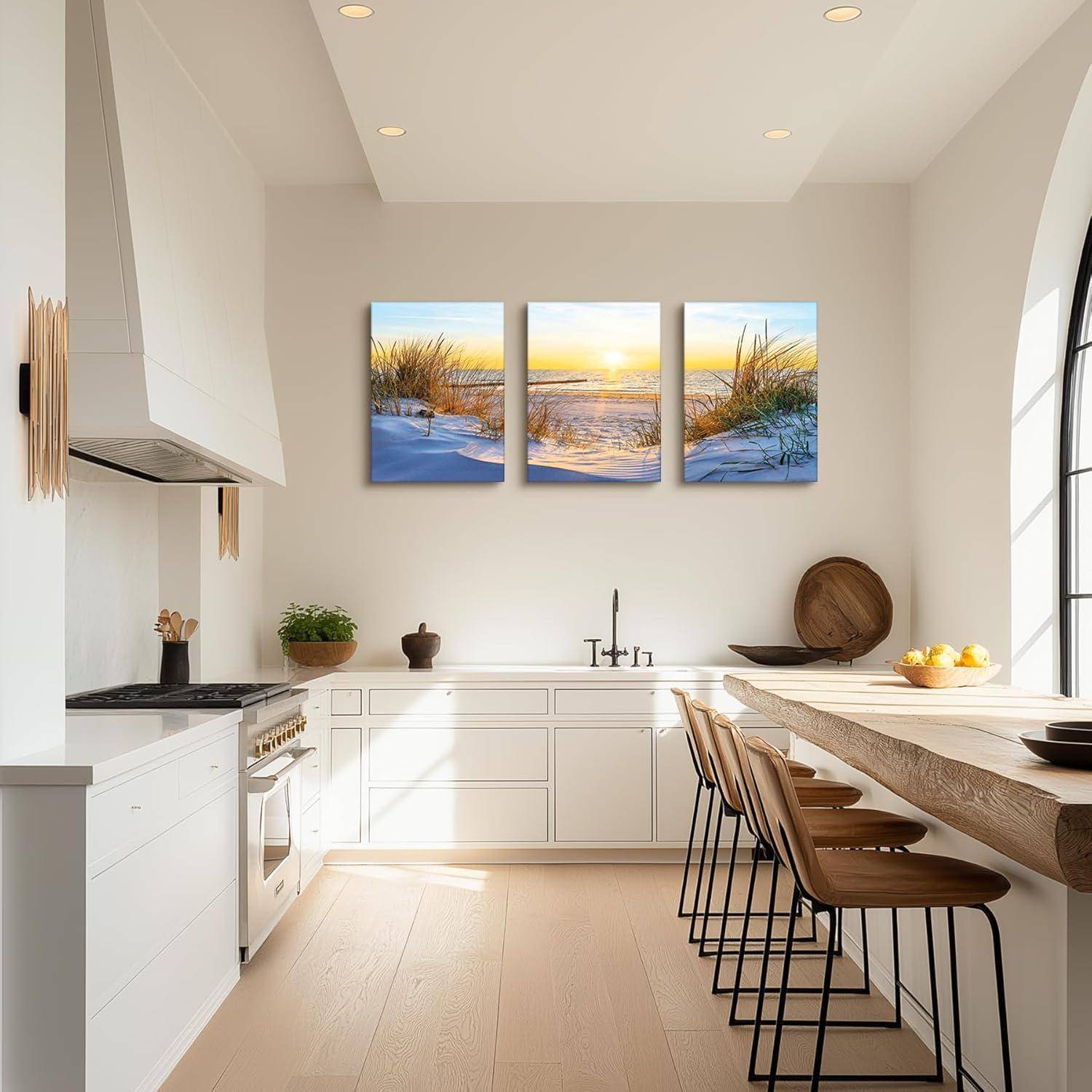 Eine moderne Küche mit weißen Schränken, einer großen Holzinsel, Barhockern und einem dreiteiligen Sonnenuntergang-Strand-Kunstwerk an der Wand. Natürliches Licht durchflutet den Raum.