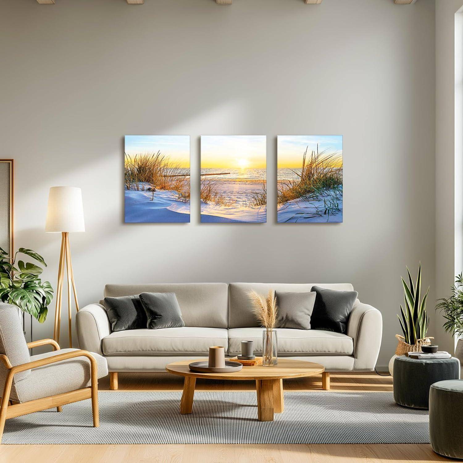 Ein moderner Wohnraum mit neutralen Tönen präsentiert ein graues Sofa, Holzmöbel, Pflanzen und ein Triptychon-Kunstwerk mit Sonnenuntergangs-Strand-Motiv an der Wand.