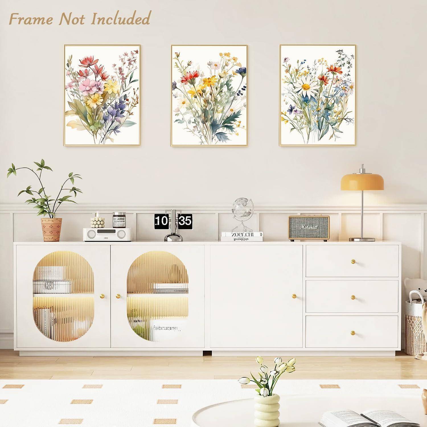 Ein moderner Wohnbereich zeigt ein weißes Sideboard mit Schubladen und Ablageregalen, dekoriert mit Vasen und Büchern. Drei Blumengemälde hängen darüber.