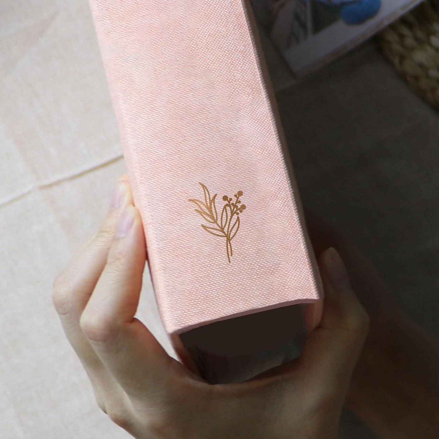 Eine Person, die ein geschlossenes rosa Buch mit goldenem Blumendesign auf dem Einband von der Seite hält, vor einem weichen beigen Hintergrund.