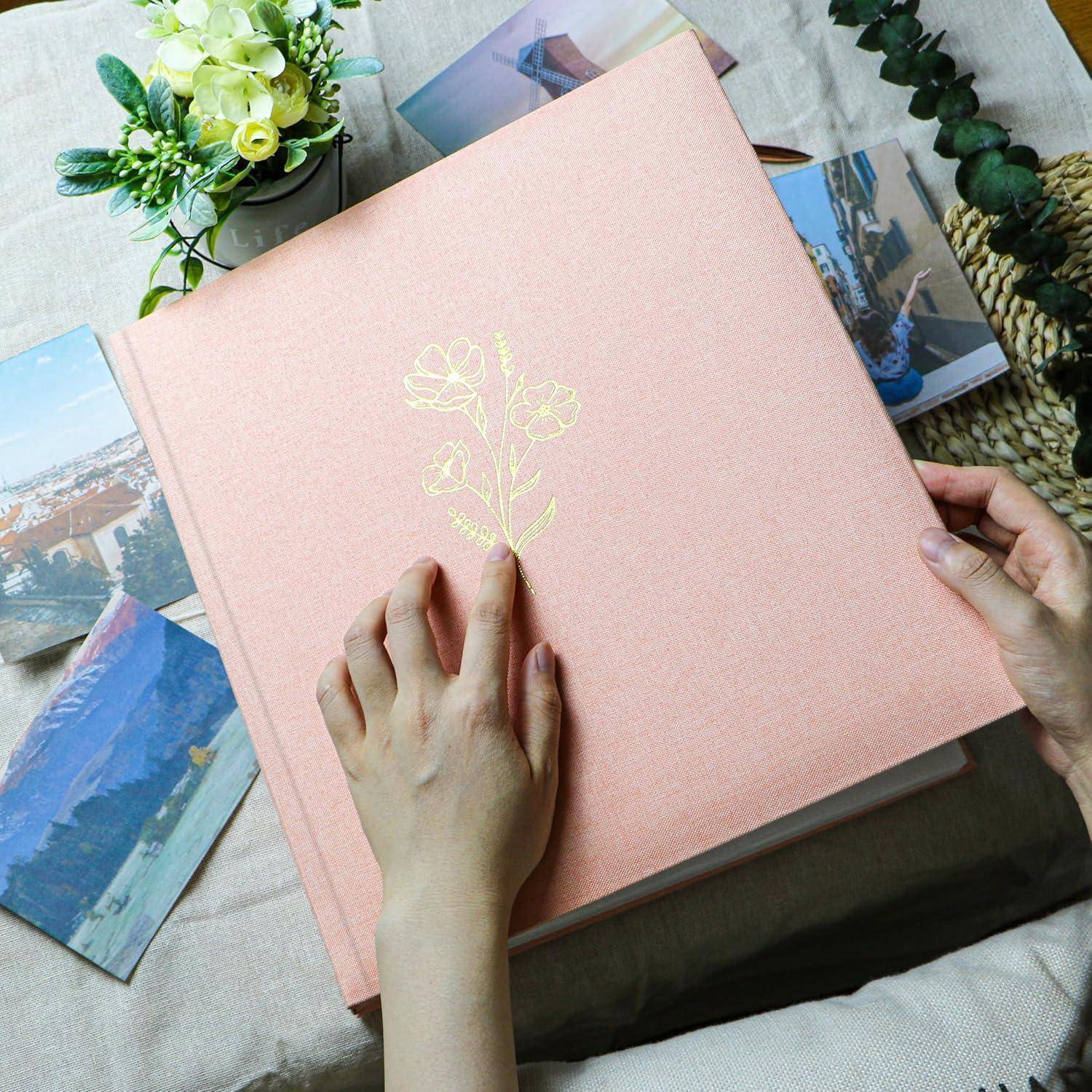 Zwei Hände halten ein rosa Fotoalbum mit einem floralen Muster auf dem Einband, umgeben von verstreuten Fotografien und einem kleinen Blumenstrauß.